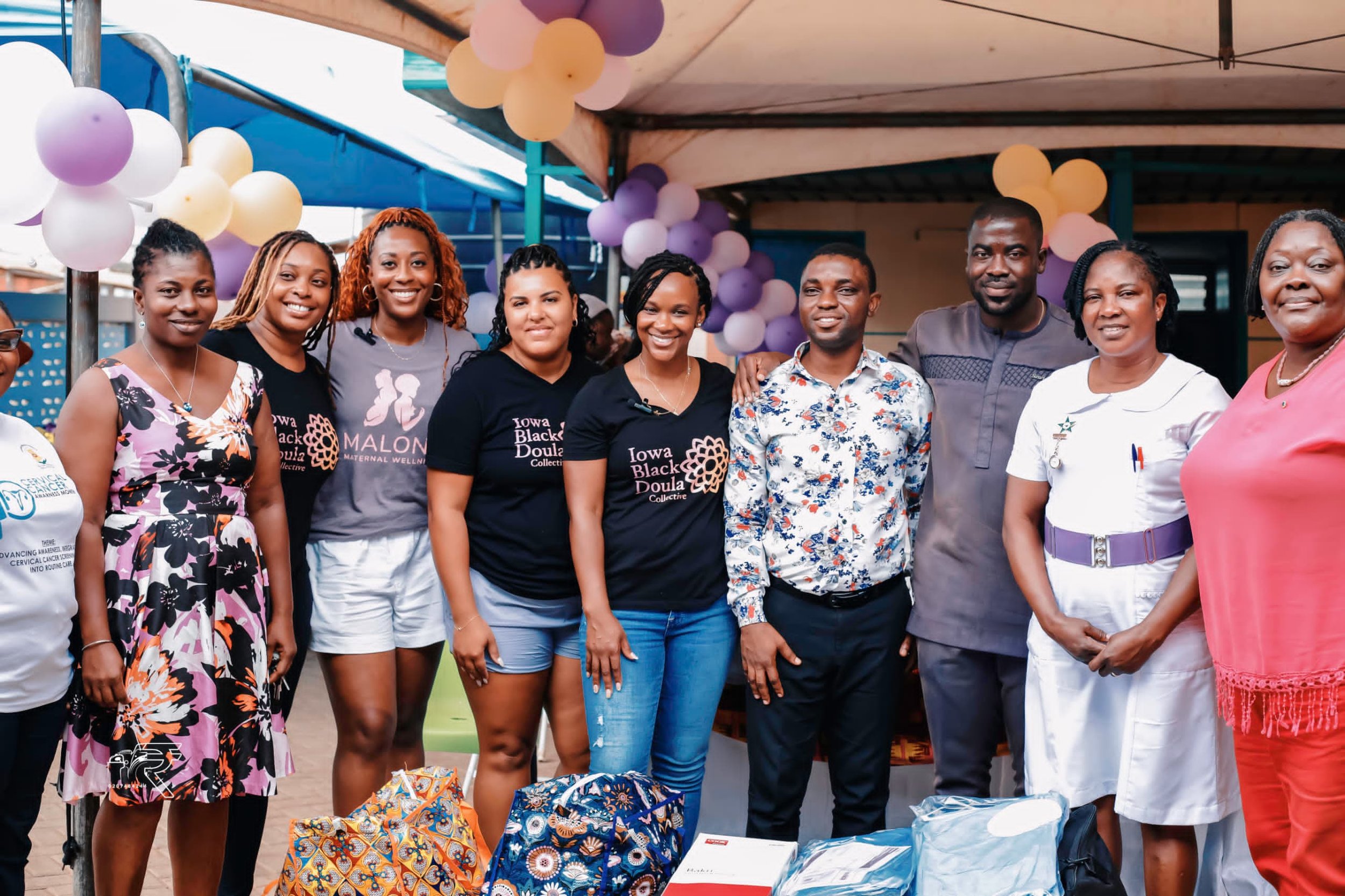 Group of diverse women and men smiling at an outdoor community event decorated with balloons, standing behind a table with various items.