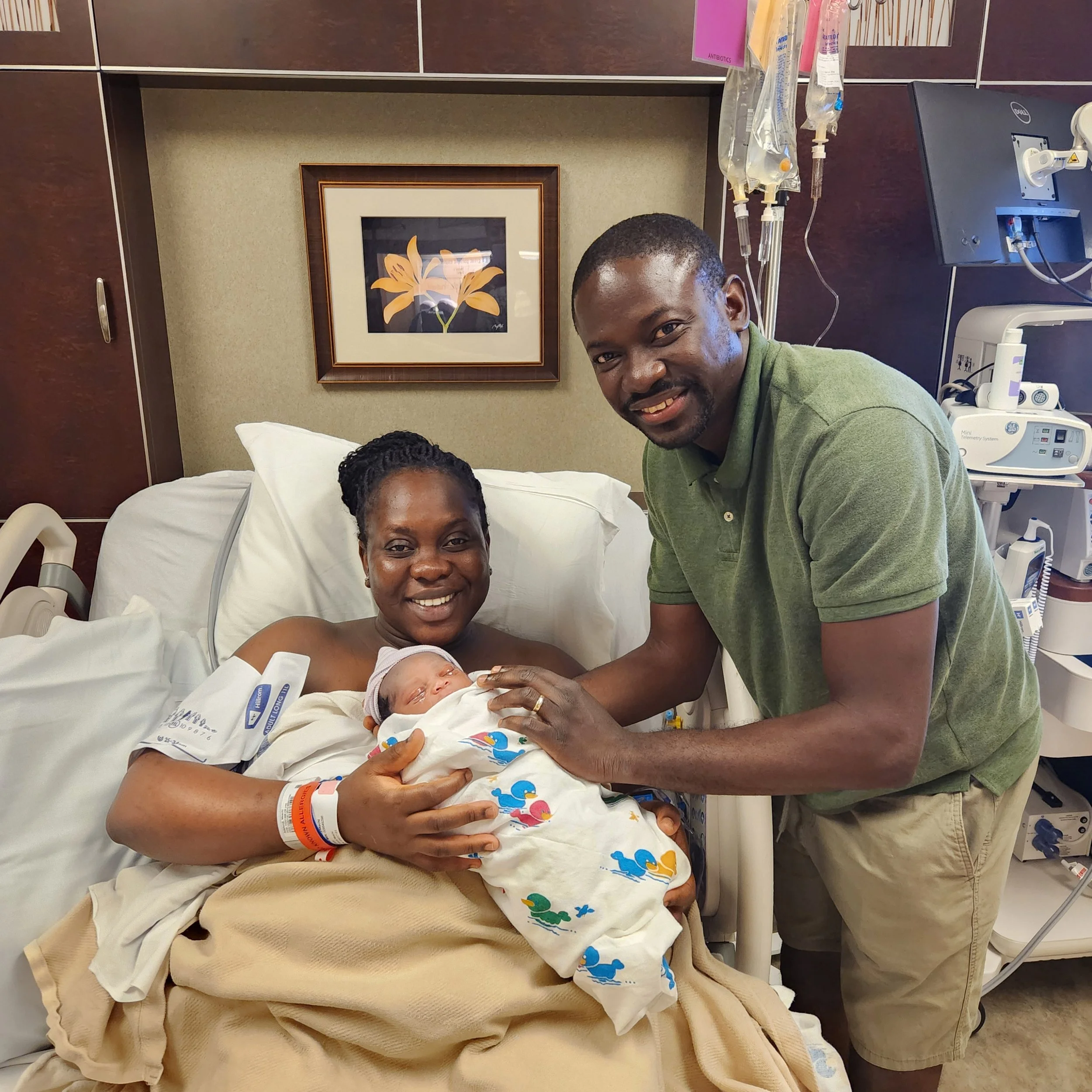 A woman lying in a hospital bed holding her newborn baby, with a man standing beside her in a hospital room. The room has medical equipment and a framed picture of a flower on the wall.