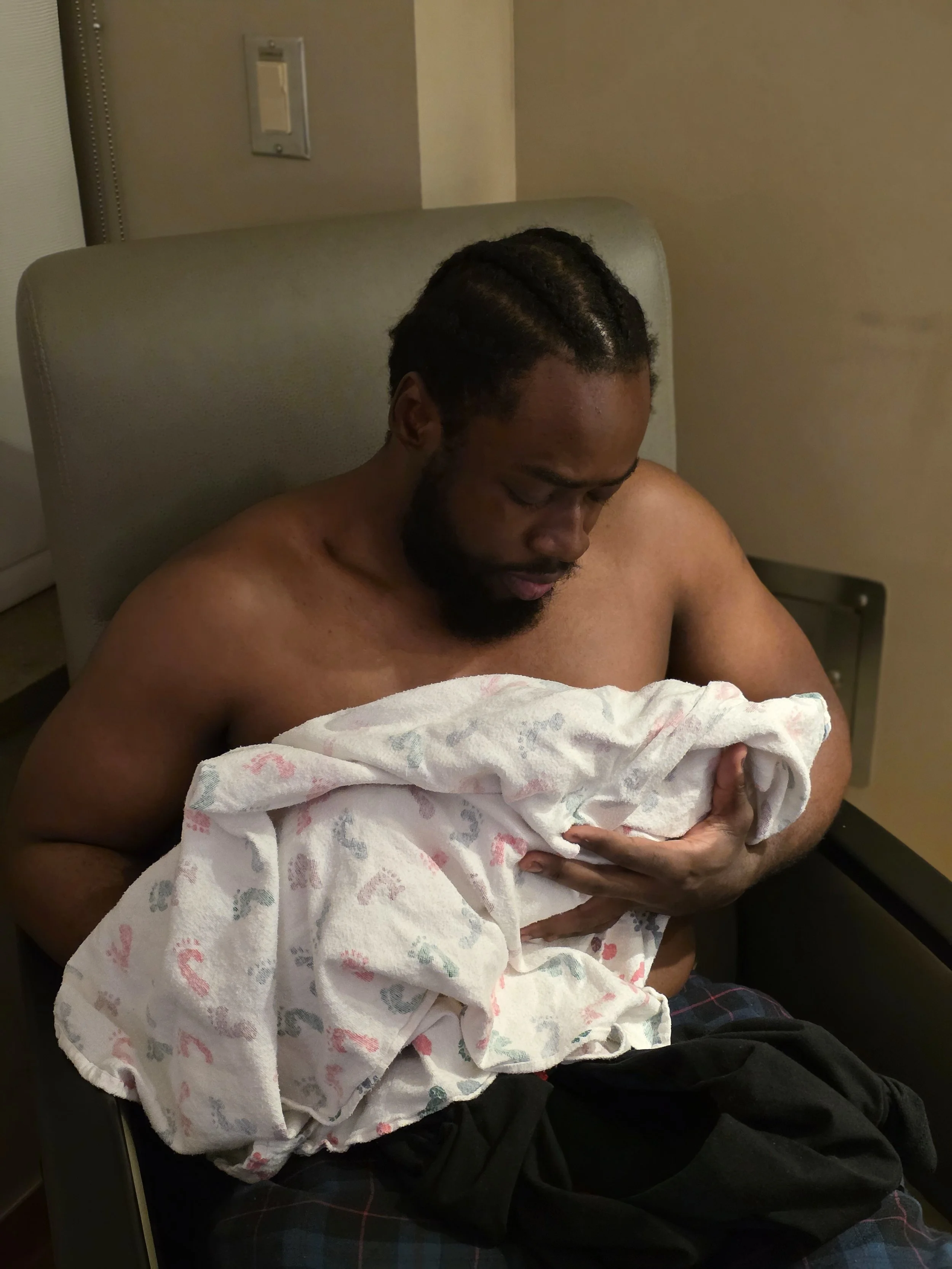 A man with braided hair and a beard is sitting in a hospital bed, holding a blanket close to his chest and looking down. The background shows typical hospital room fixtures like a light switch.