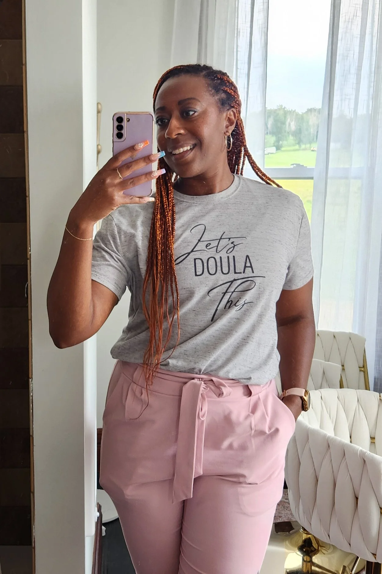 Self-portrait of a woman taking a mirror selfie indoors, dressed in a gray t-shirt that reads 'Let's Doula This' and pink pants tied with a bow, with a bright window behind her showing a green outdoor landscape.