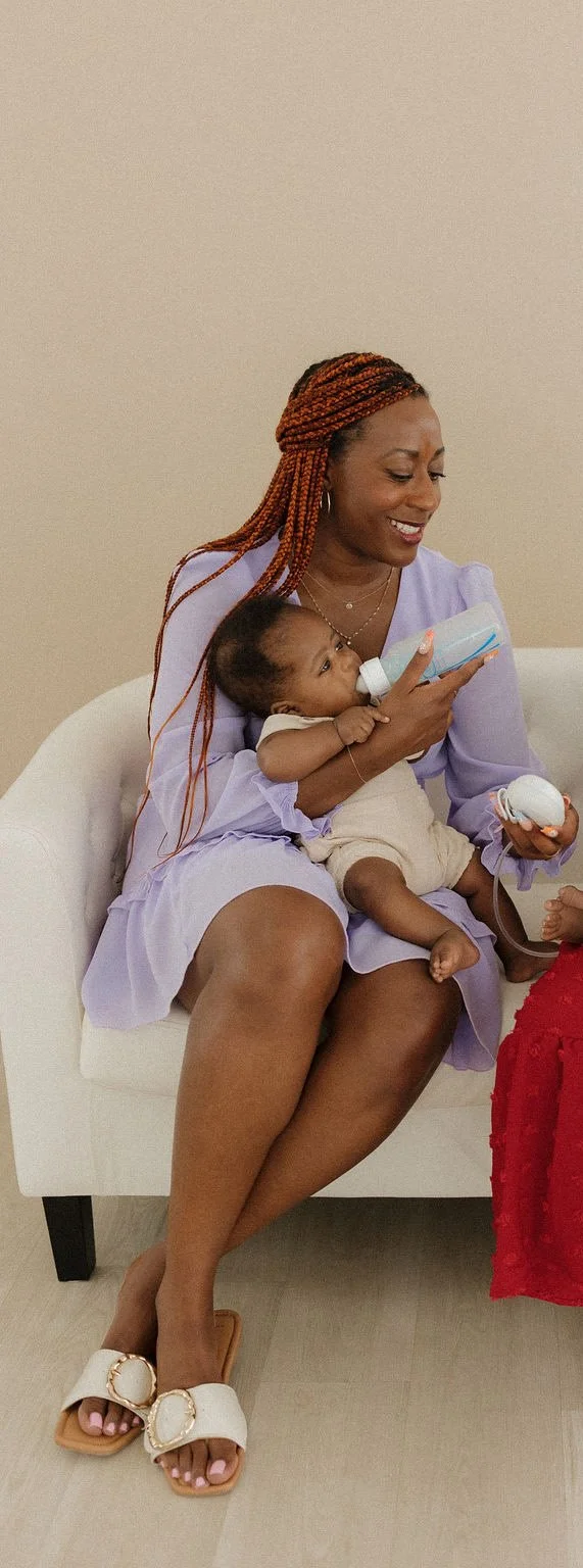 Woman with braided hair feeding a baby with a bottle while sitting on a white couch.
