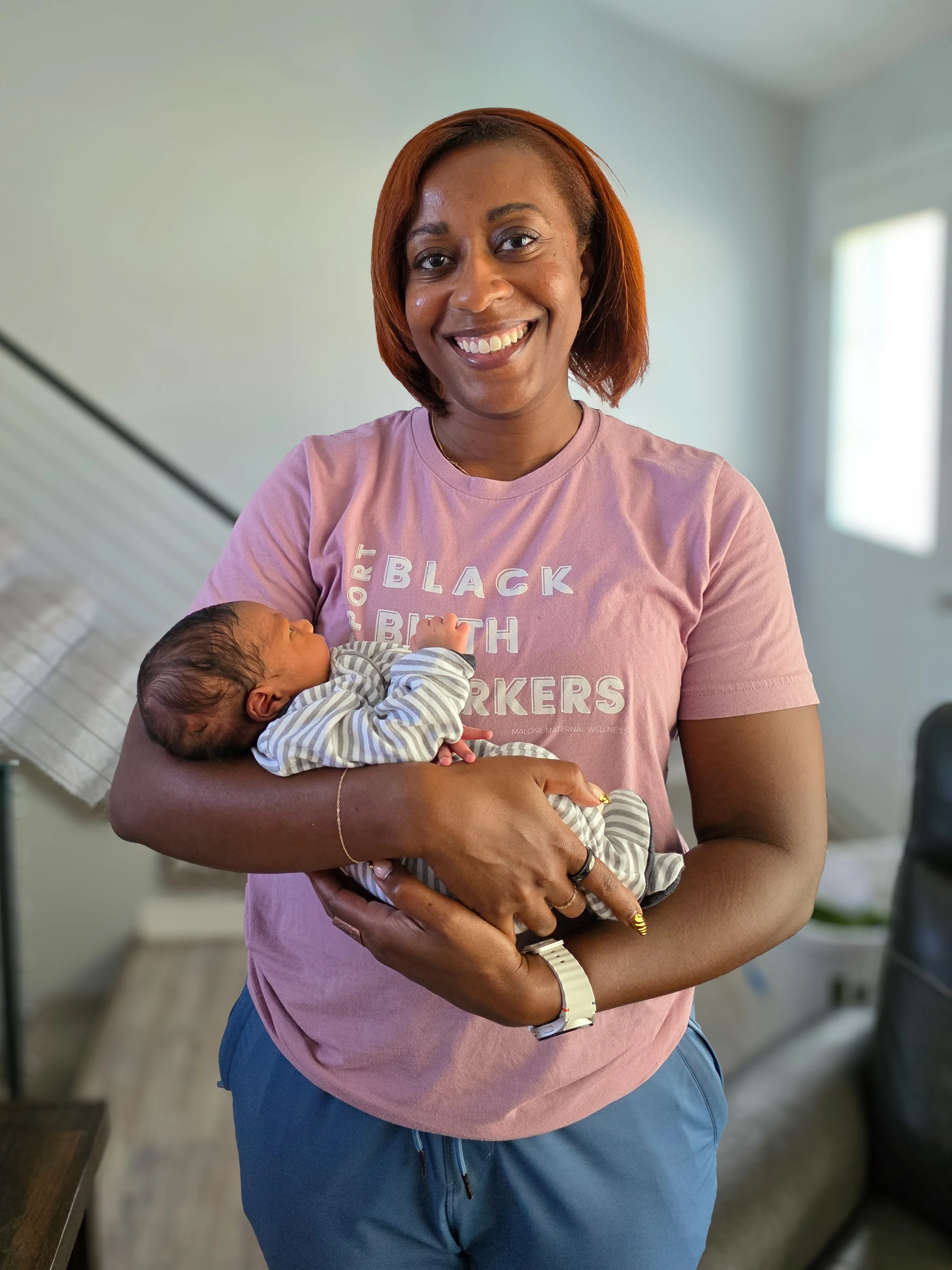 A woman holding a newborn baby indoors during the daytime. She is wearing a light pink T-shirt and blue pants, smiling at the camera. The background features a decorated staircase and a window letting in natural light.