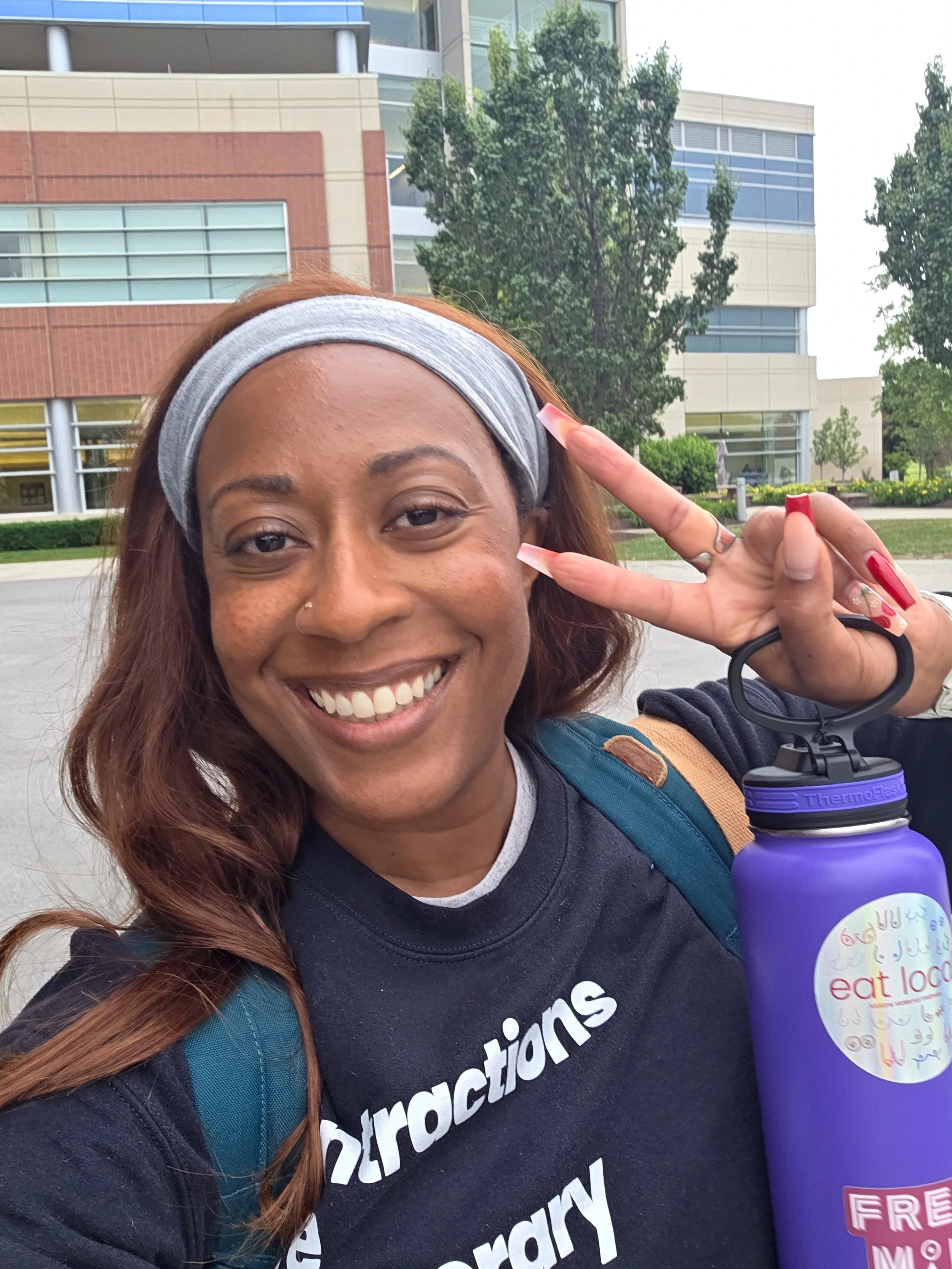 A woman with reddish-brown hair, wearing a gray headband and black t-shirt with white text, smiling and holding a purple water bottle in front of a modern building with trees and greenery.