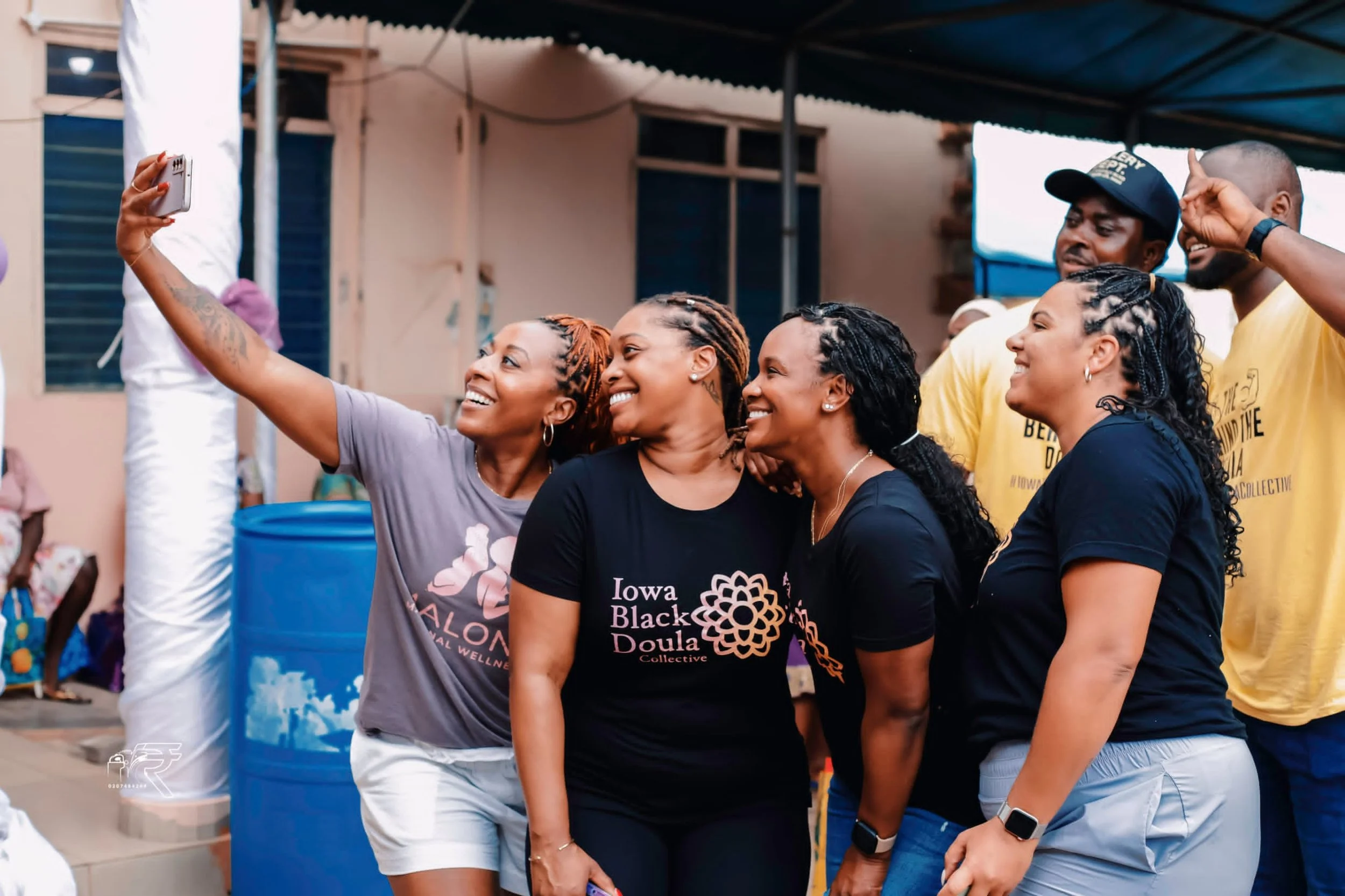 Group of smiling women and men taking a selfie at an outdoor event, with some wearing black shirts that say 'Iowa Black Doula Collective' and others wearing yellow T-shirts. They are standing near a building with a blue water barrel nearby.