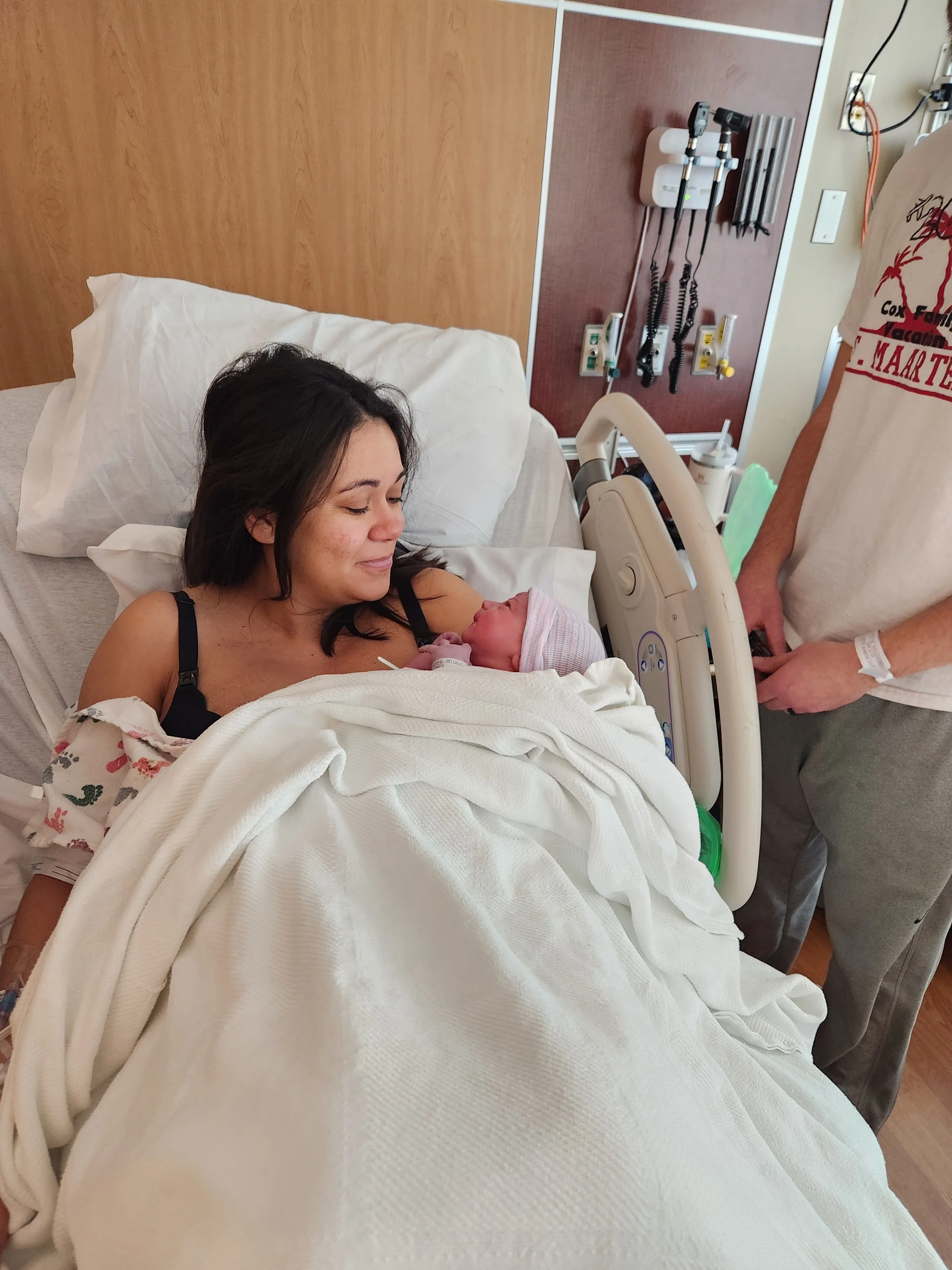 A woman lying in a hospital bed, holding a newborn baby wrapped in a blanket, with medical equipment and a nurse or doctor standing nearby.