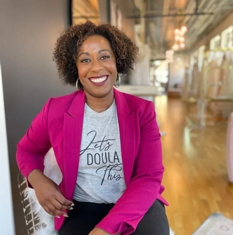 A woman smiling, wearing a hot pink blazer and a light gray T-shirt with the text 'Let's Doula This' in black cursive, sitting in a bright, modern indoor space with bookshelves and large windows.