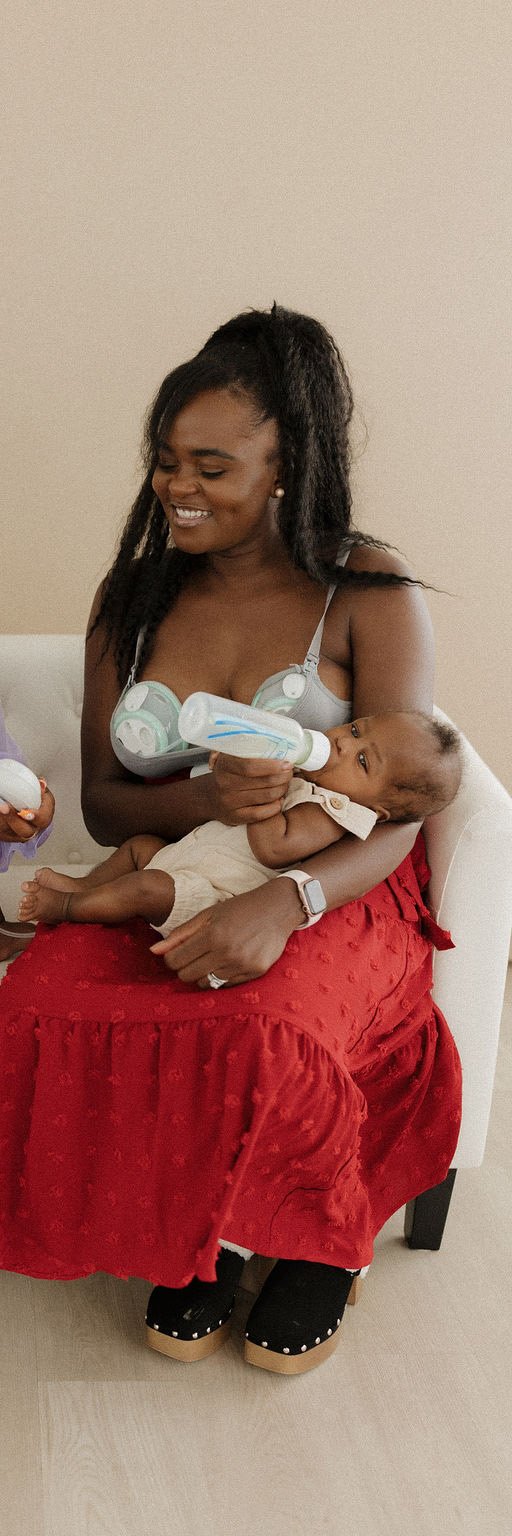 A woman sitting on a white chair, feeding a baby with a bottle. The woman is smiling and wearing a red skirt, a gray top with a baby monitor, and slippers. The baby is lying across her lap, looking at the bottle.