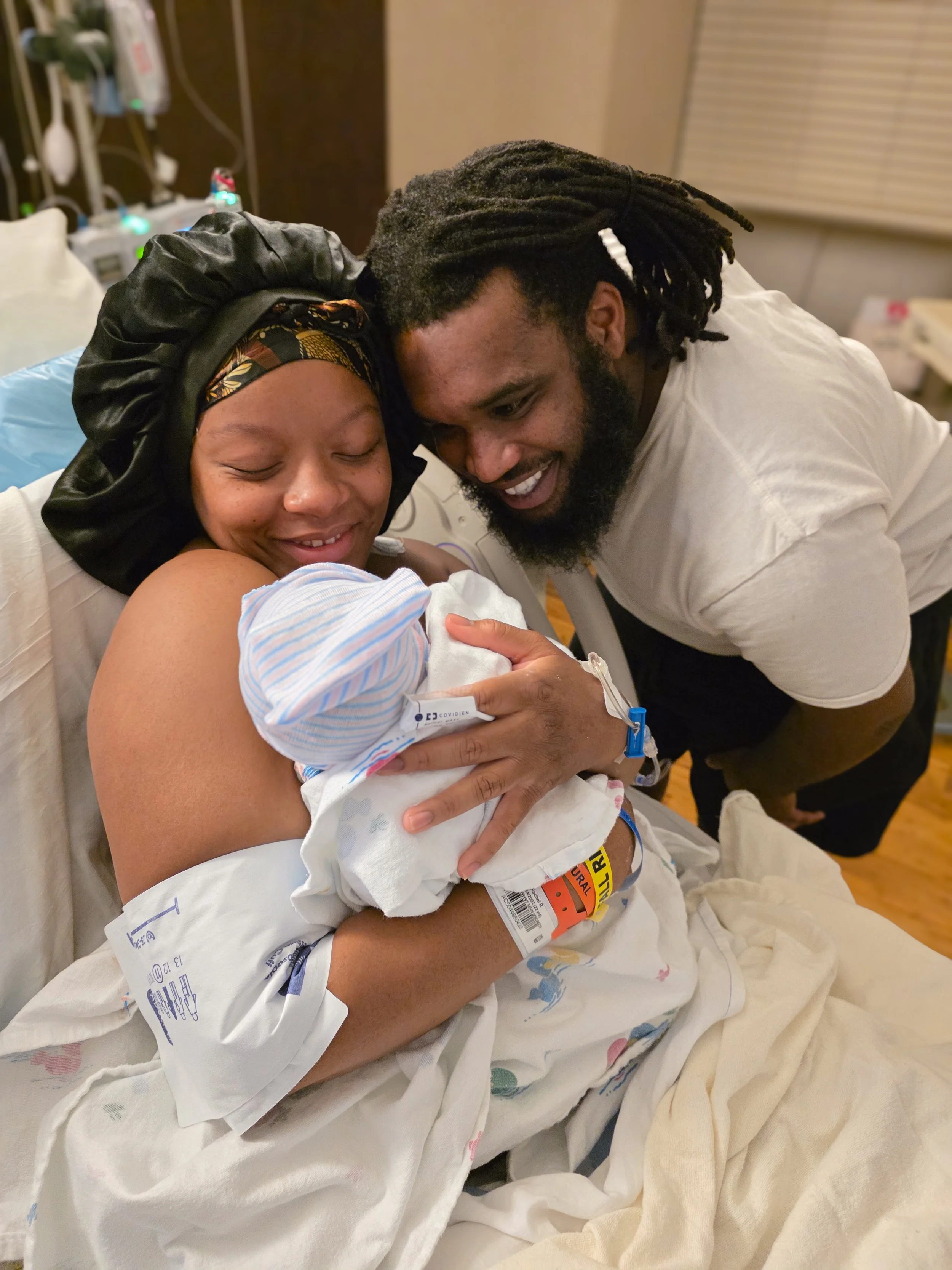 A woman in a hospital bed holding a newborn baby, with a man leaning over her, all smiling and looking happy.