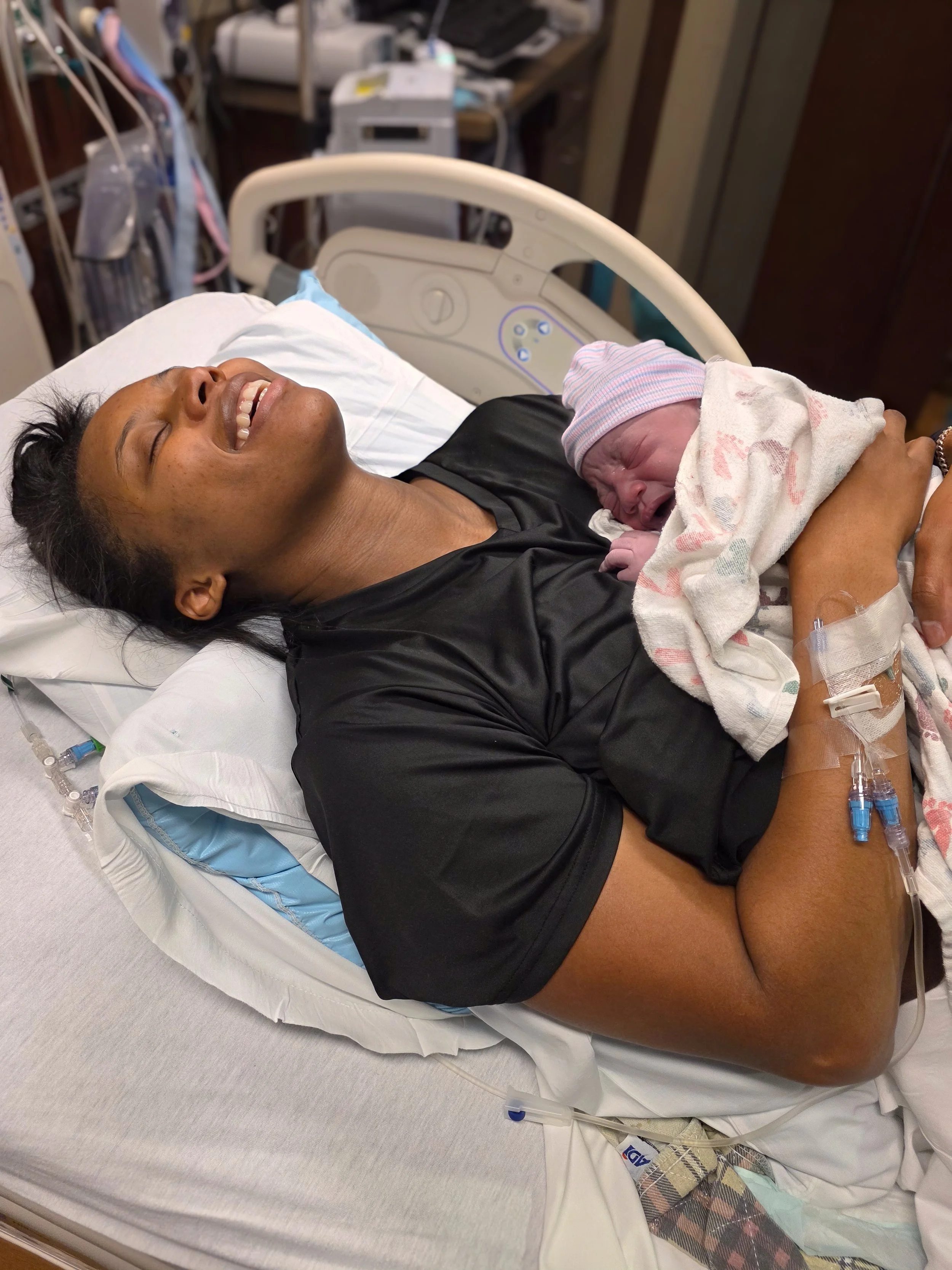 A woman in a hospital bed holding a newborn baby wrapped in a blanket, both smiling and happy.