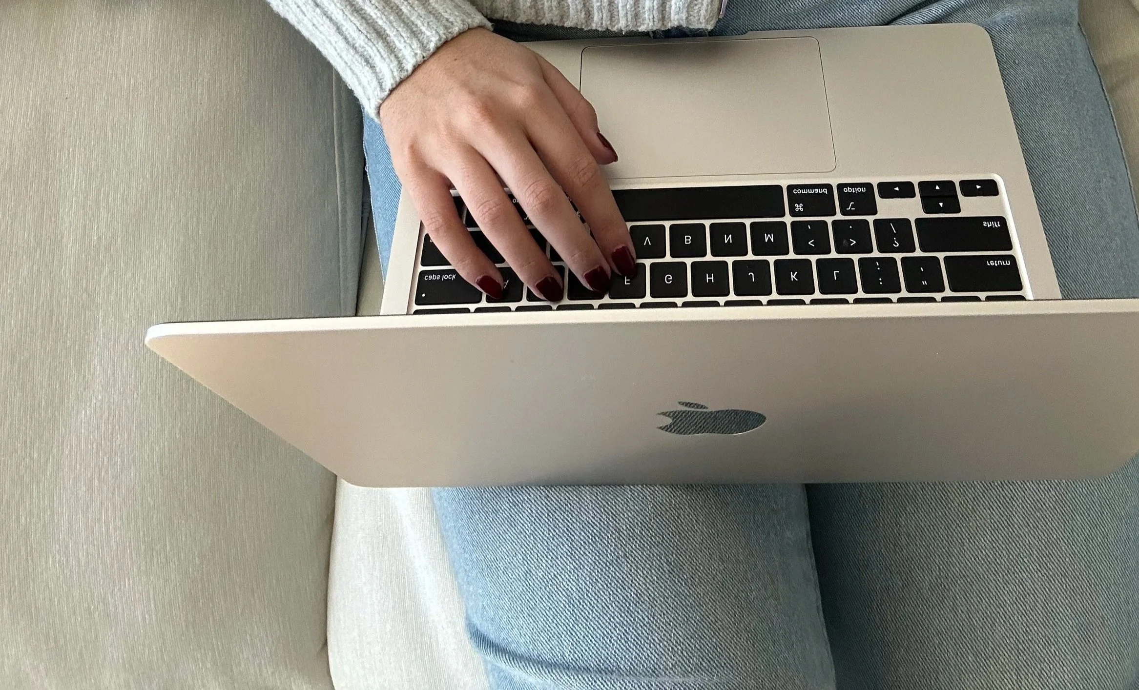 A person with painted nails typing on a silver MacBook laptop on a blue and gray striped couch.