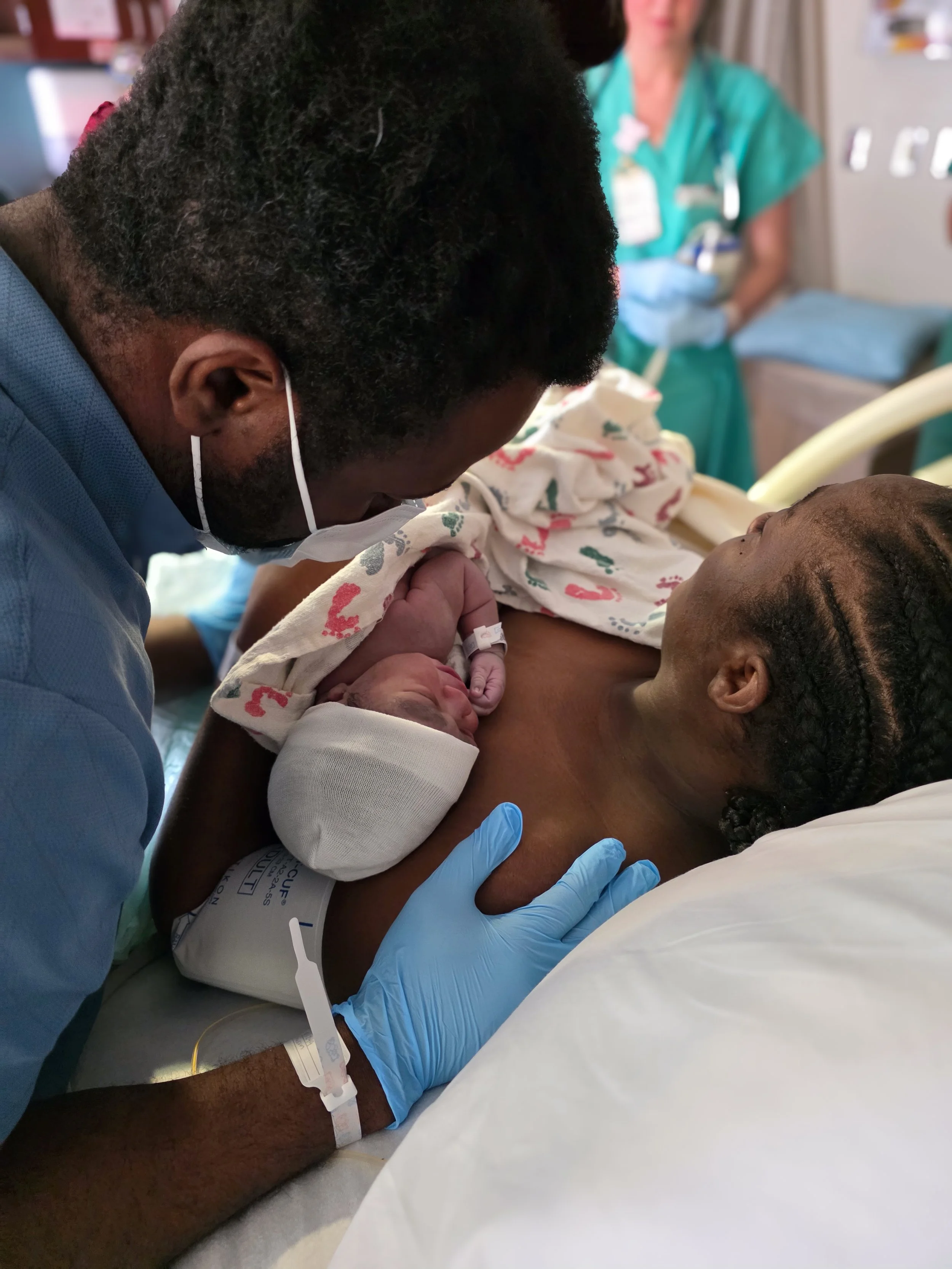 A newborn baby is held by a woman lying on a hospital bed. A healthcare professional in teal scrubs and gloves is observing nearby.