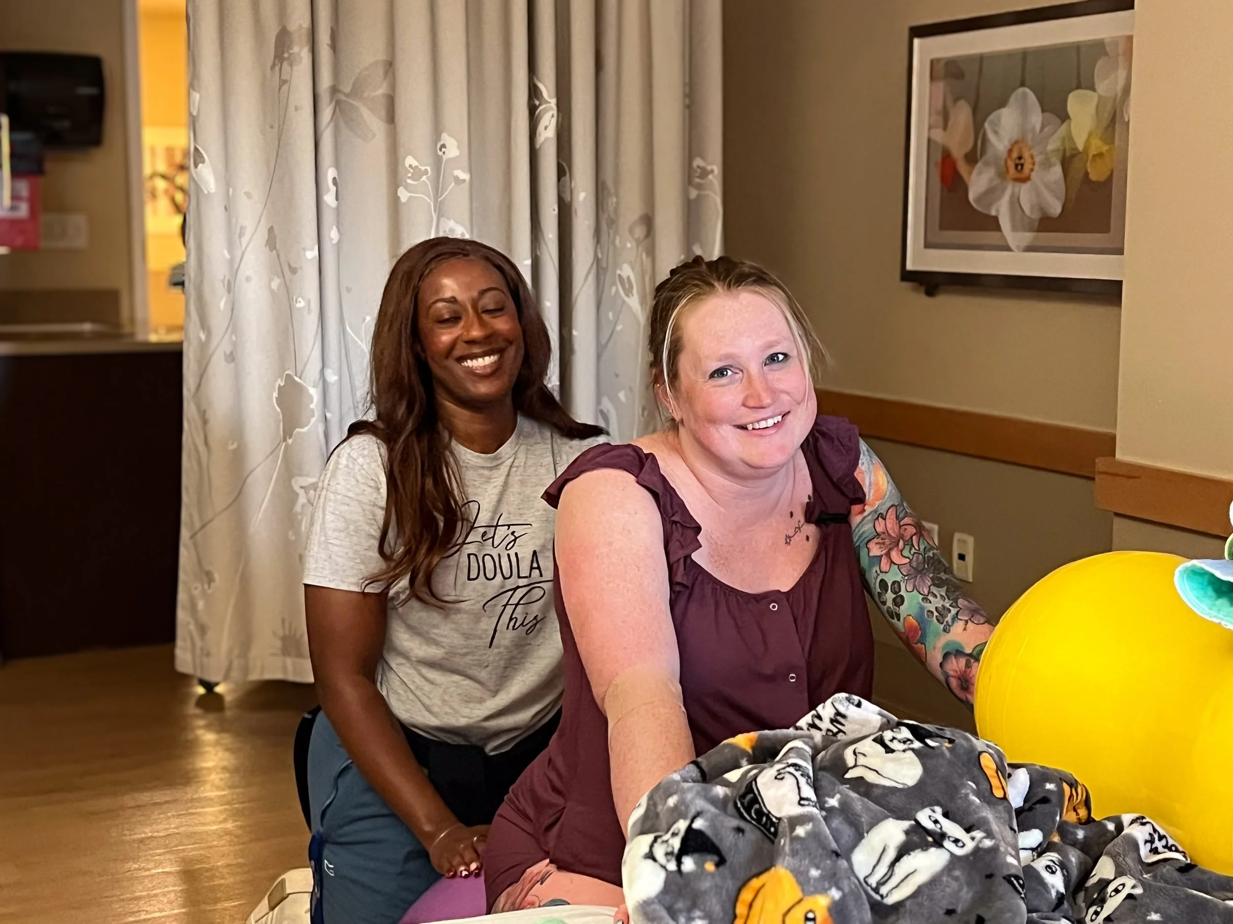 Two women, one with long dark hair and the other with short blonde hair, are sitting at a hospital bed smiling. The blonde woman is in front, and the dark-haired woman is behind her.