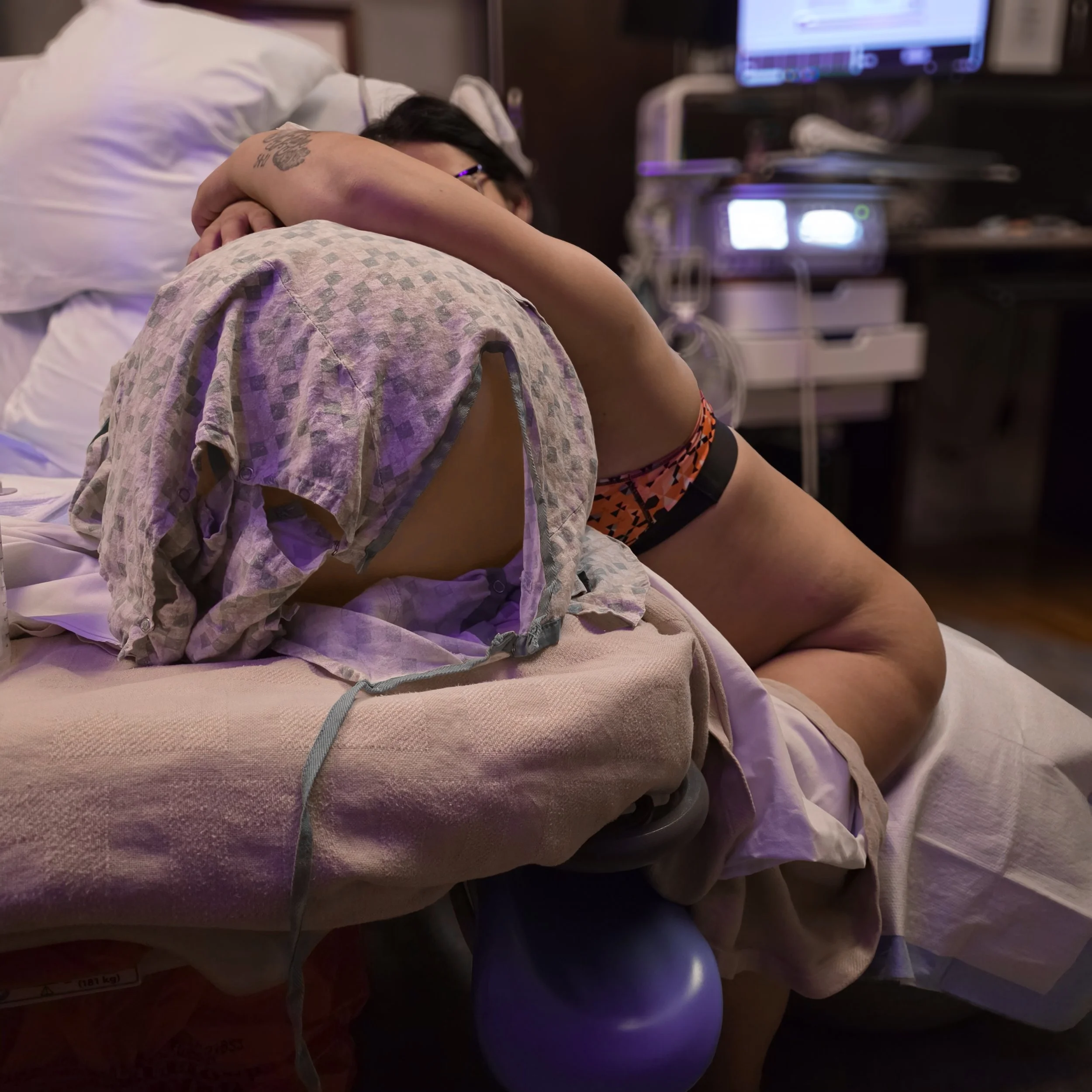 A woman with dark hair, glasses, and a tattoo on her left arm lying in a hospital bed, resting her head on a pillow, with medical equipment and a monitor in the background.