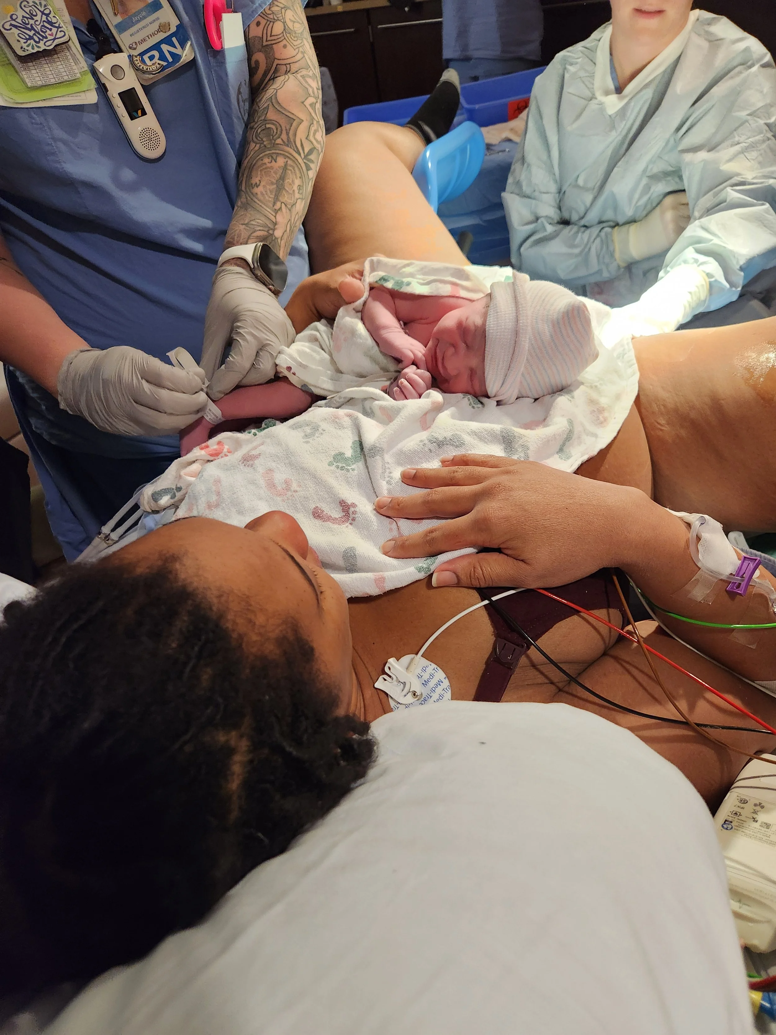 A newborn baby wearing a hospital cap and wrapped in a blanket, lying in a hospital bed with a woman lying beside them, a healthcare worker in scrubs and a tattooed arm assisting, and a person using a phone with monitors and medical equipment nearby.