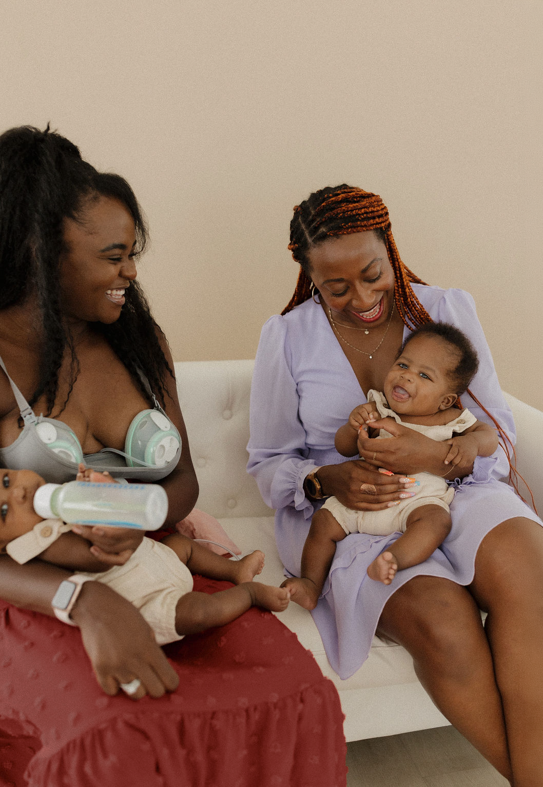Two women and two babies sitting on a beige couch, smiling and interacting with each other. One woman is holding a baby, and the other woman is wearing a tank top with ear protection around her neck.