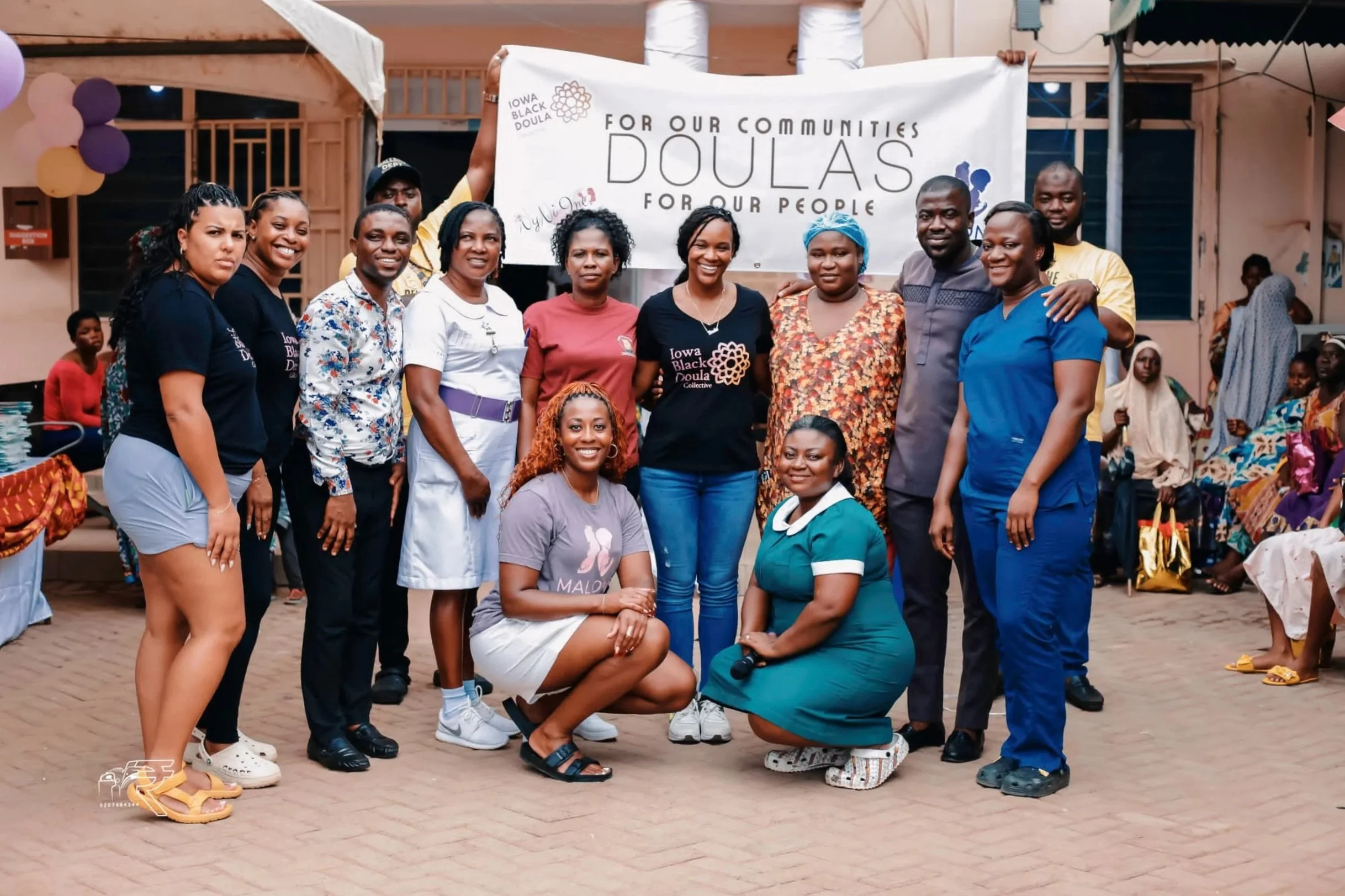 Group of people standing together outdoors, smiling for the camera, with a banner in the background that reads 'For Our Communities, Doula, For Our People.' Some are wearing t-shirts with the same logo, and there are additional people and decorations in the background suggesting a community event or gathering.
