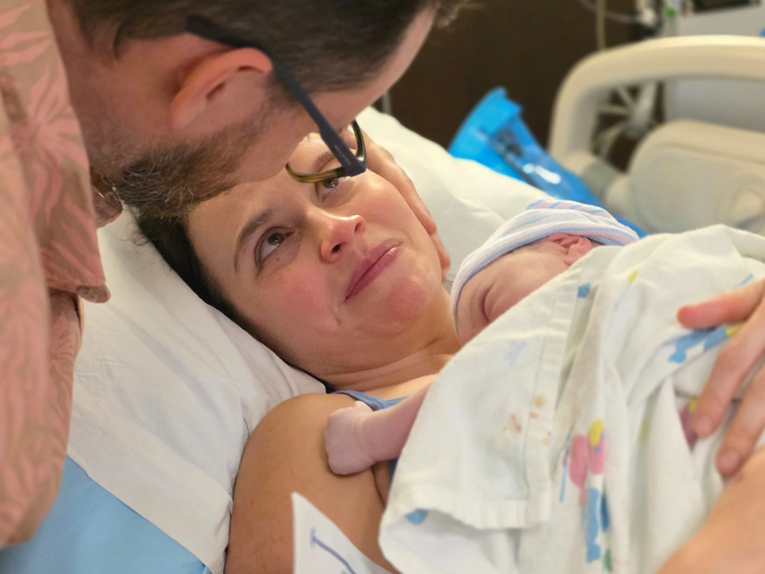 A woman lying in a hospital bed holding her newborn baby, with a man leaning over her, visible wearing glasses and a beard, in a hospital room.