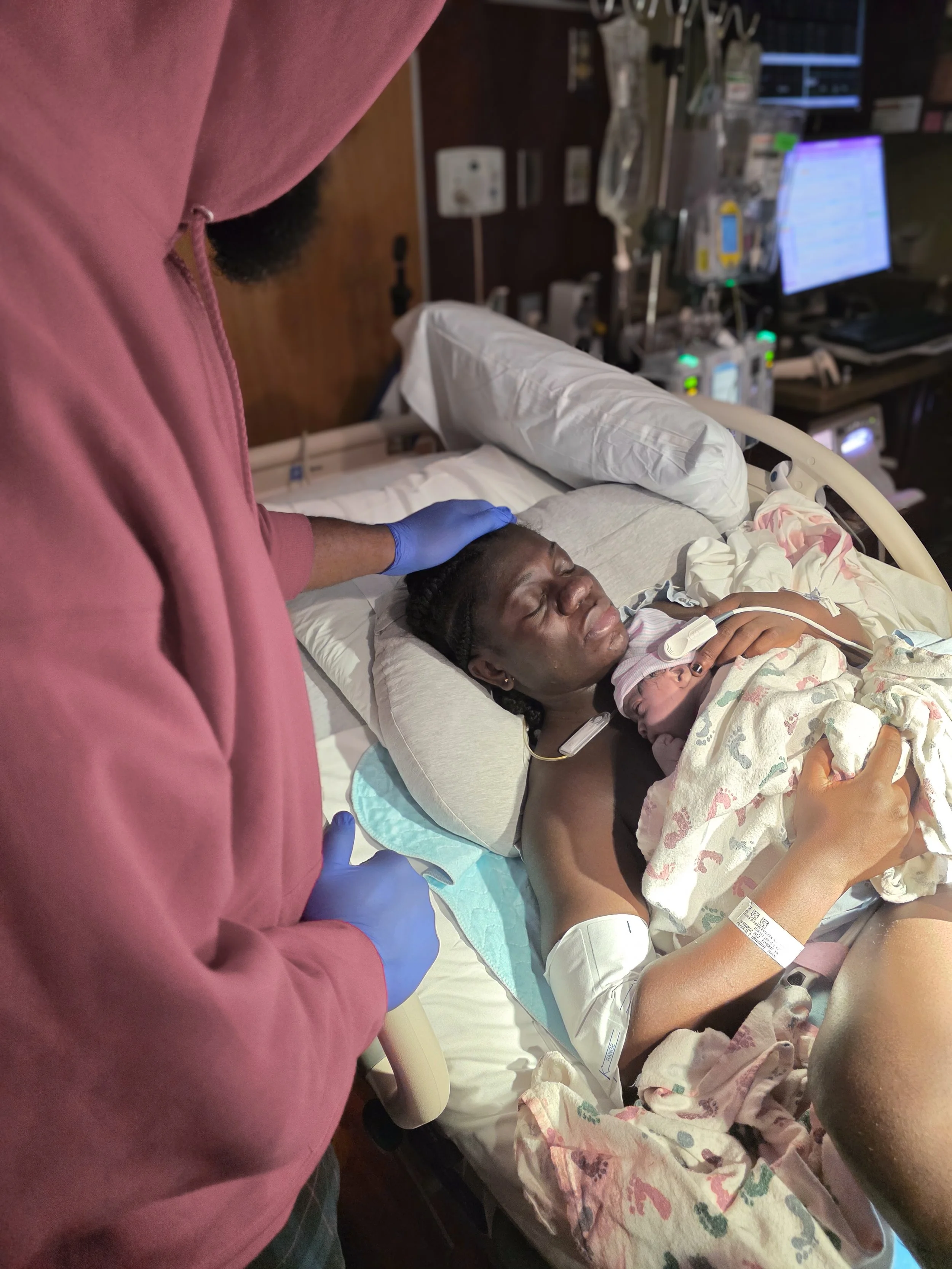 A woman is lying in a hospital bed, holding a newborn baby close to her chest. A person in a pink hoodie and blue gloves is beside her, attending to her. Medical monitors and equipment are visible in the background.