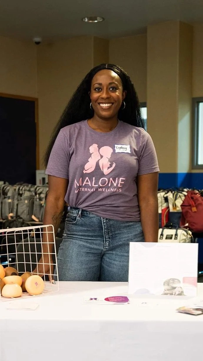 A woman standing behind a table at a wellness event, wearing a purple T-shirt that reads 'Malone Maternity Wellness' and a handwritten name tag, smiling at the camera.