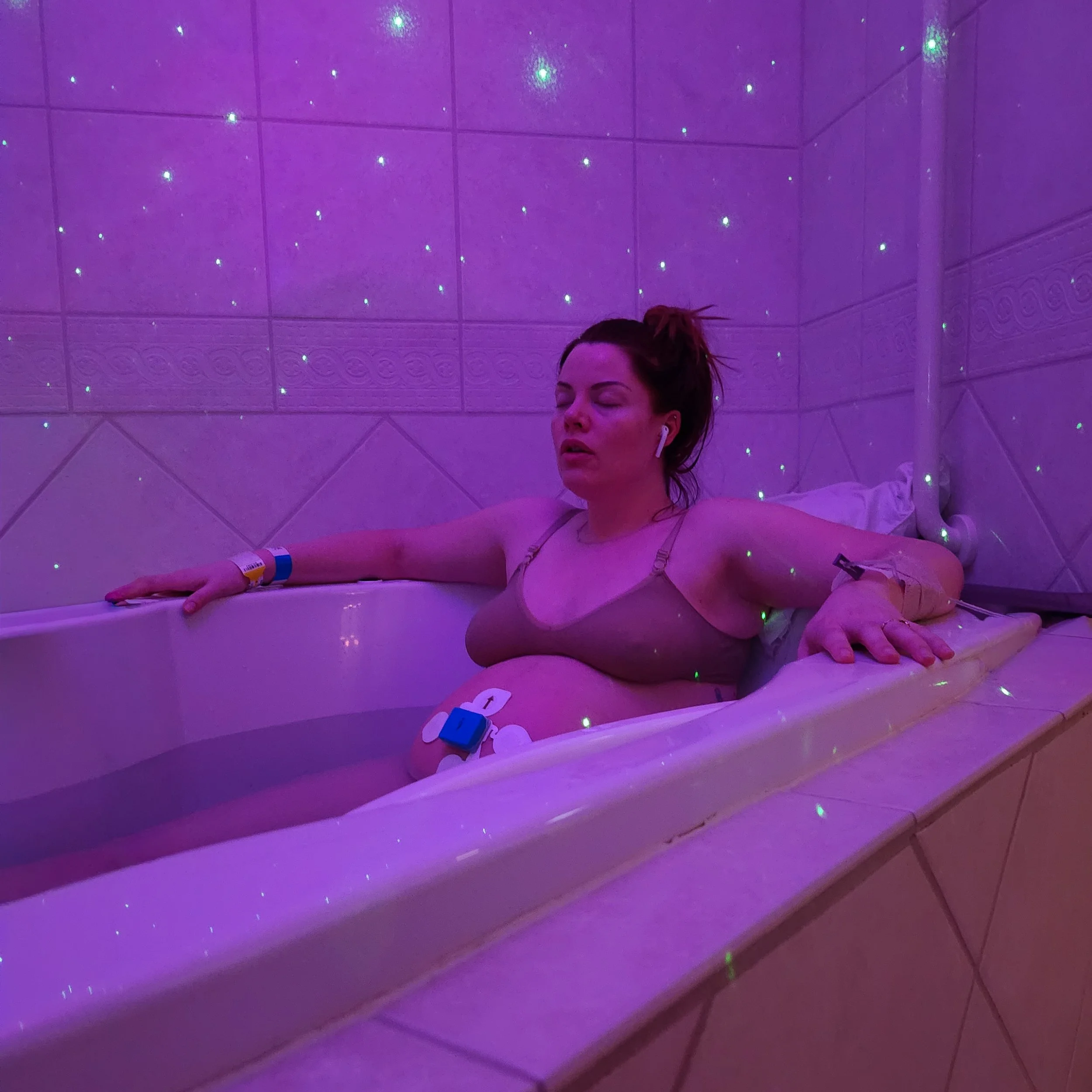 A woman with closed eyes and AirPods in her ears is relaxing in a pink-lit room with starry projector lights on the ceiling.