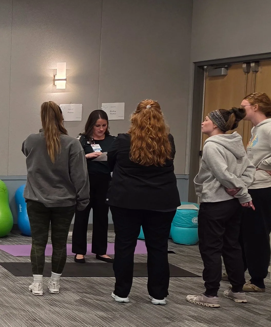 Group of five women standing indoors, casually dressed, engaged in conversation with a woman in a black uniform showing something on a phone. There are exercise mats and fitness equipment in the background.