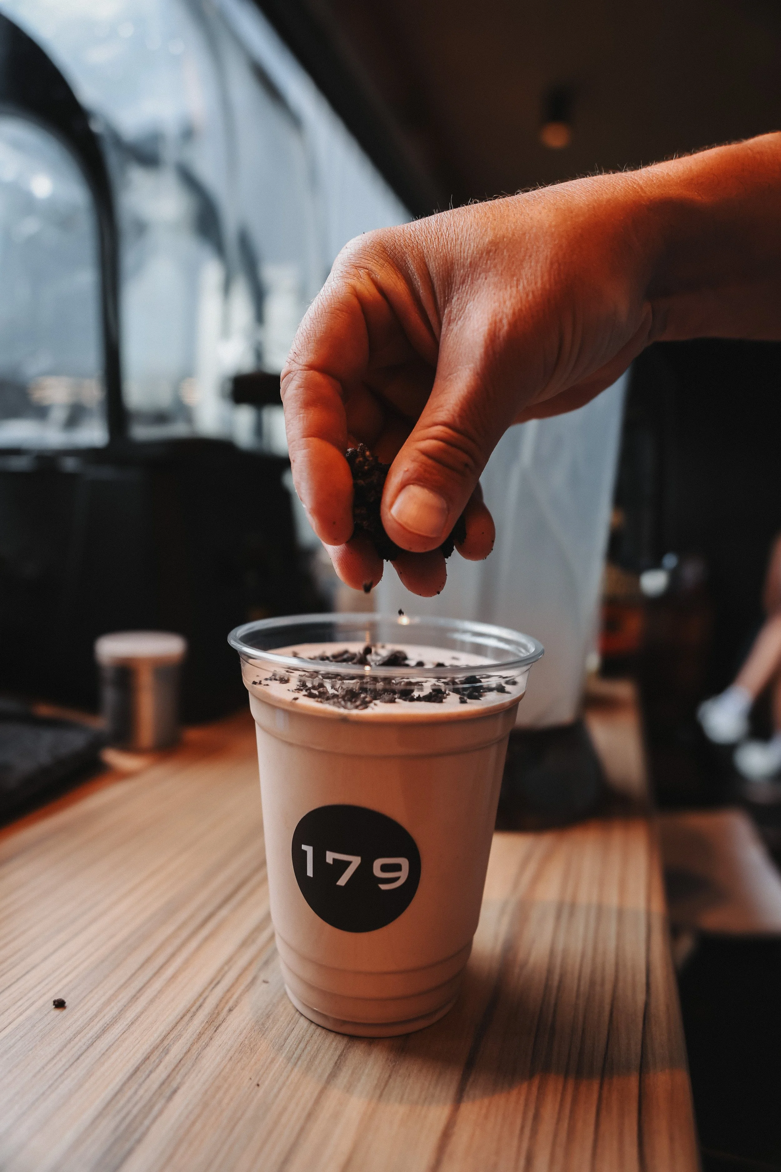 A hand is sprinkling chocolate shavings on a tea-colored beverage in a paper cup with a black circle and white number 179 printed on it. The cup is placed on a wooden table.
