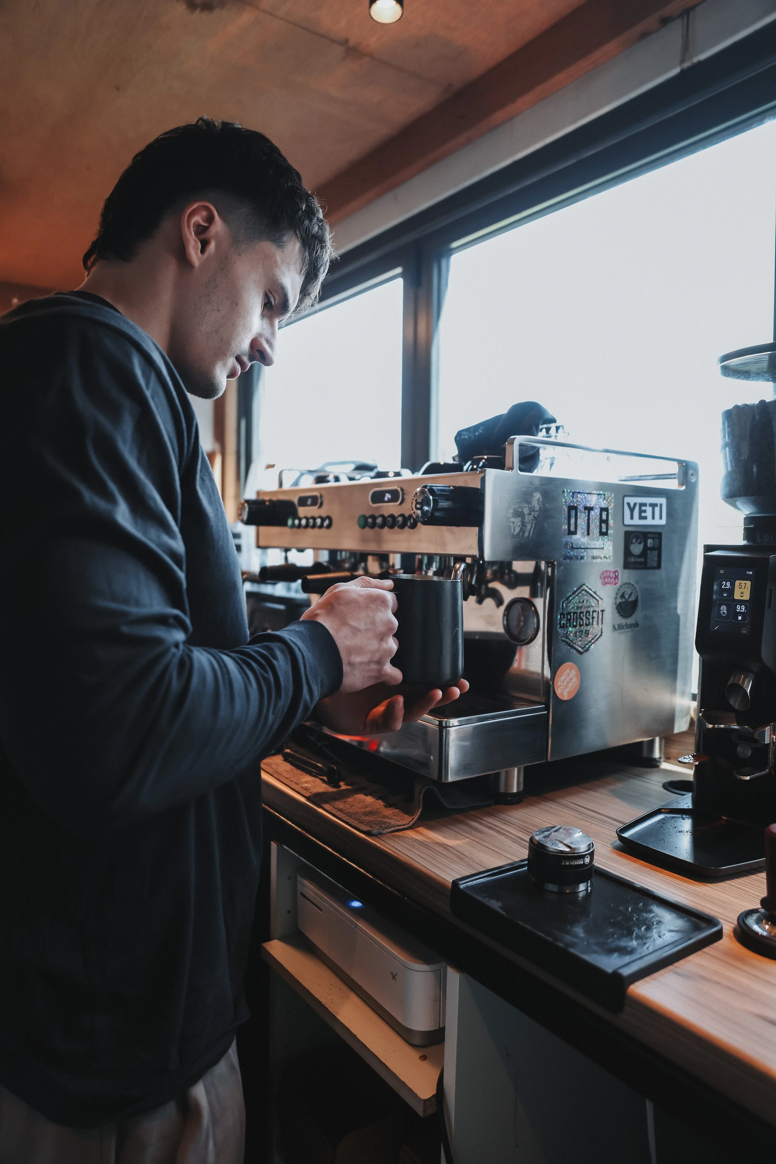 A young man wearing a black long-sleeve shirt preparing coffee using an espresso machine in a cozy kitchen with wooden accents and large windows.