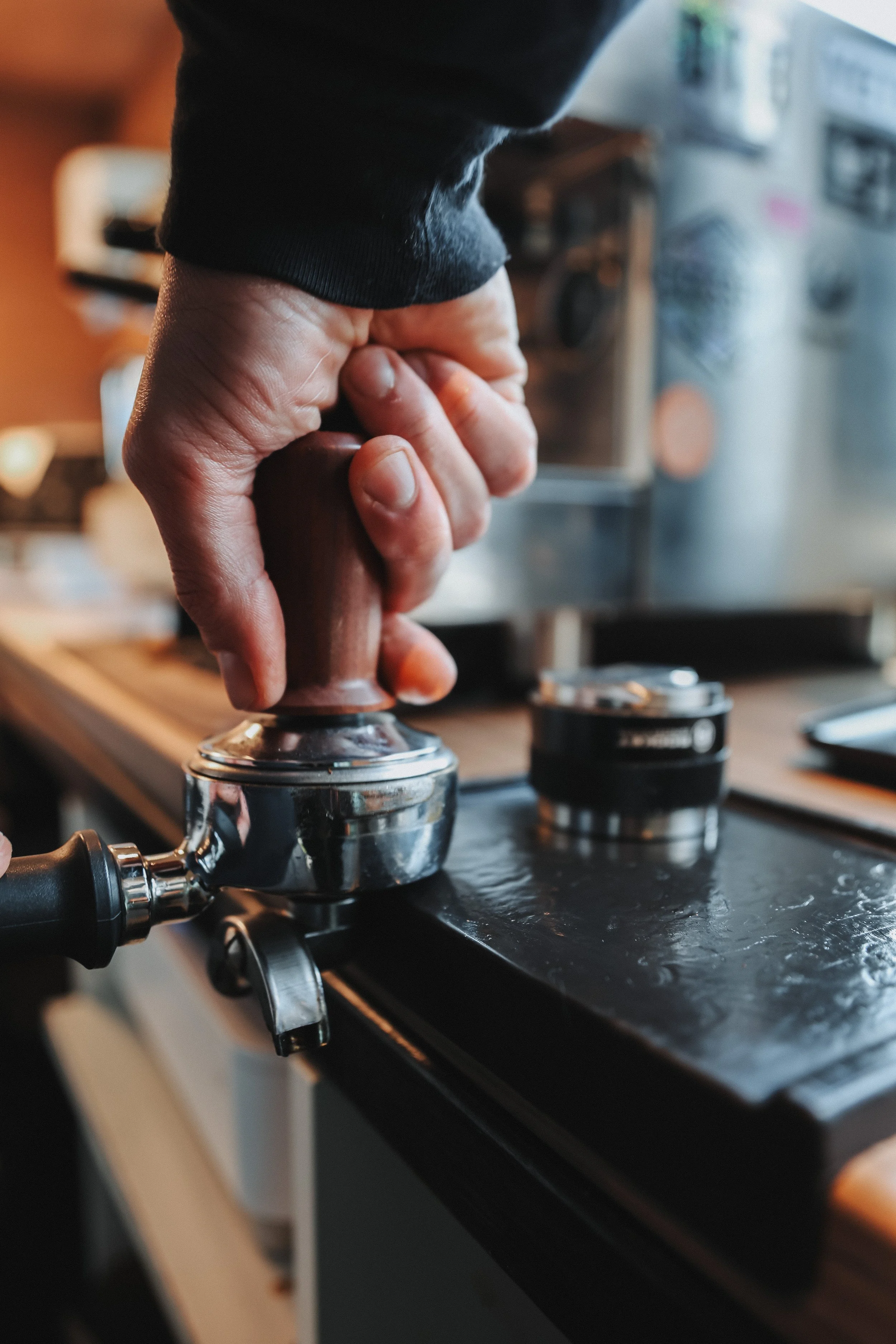 A person pressing down on a metal espresso tamper with one hand, preparing to tamp coffee grounds in a portafilter on a black countertop with espresso machine components visible.