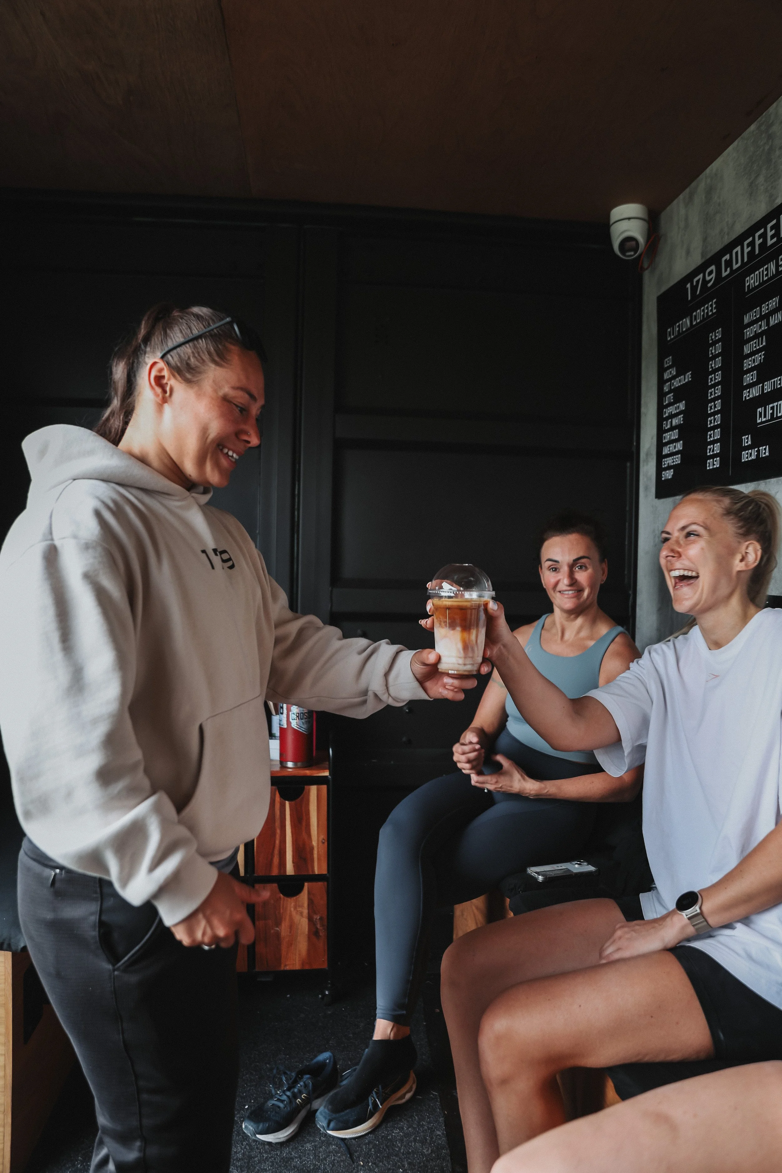 A woman with glasses on her head is smiling and handing a beverage in a plastic cup to a woman wearing a white shirt, sitting on a bench, in a coffee shop. Two other women are sitting and smiling in the background.
