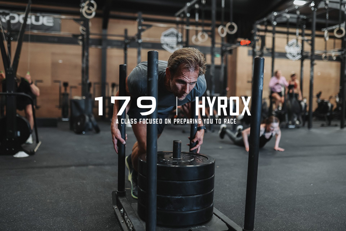 Man pushing a weighted sled at a gym, with other people working out in the background.