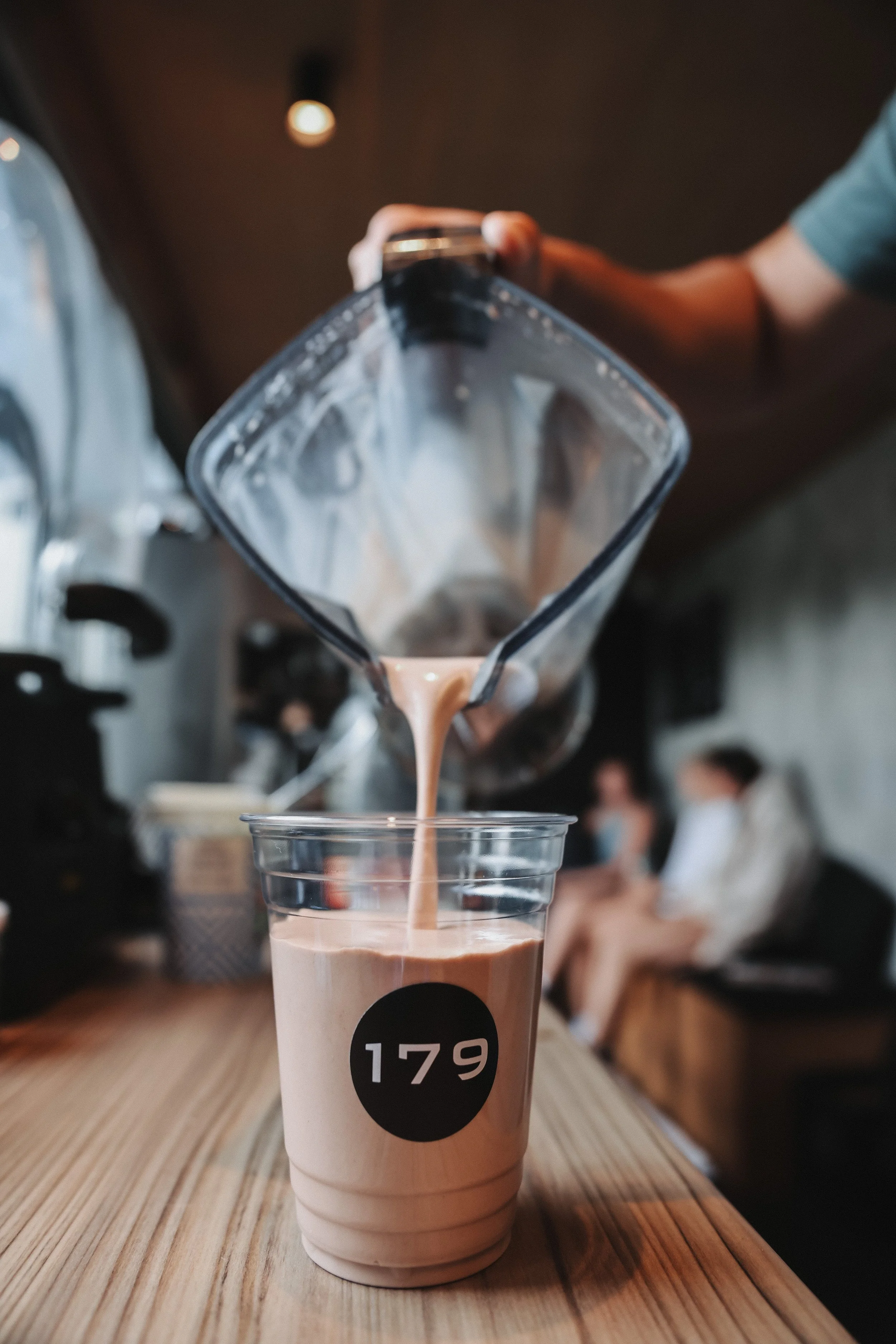 A hand pouring a chocolate milkshake from a blender into a clear plastic cup with a black circle and white number 179 on it. The drink has a swirl of chocolate on top. The scene appears to be in a cafe or restaurant with blurred background of people.
