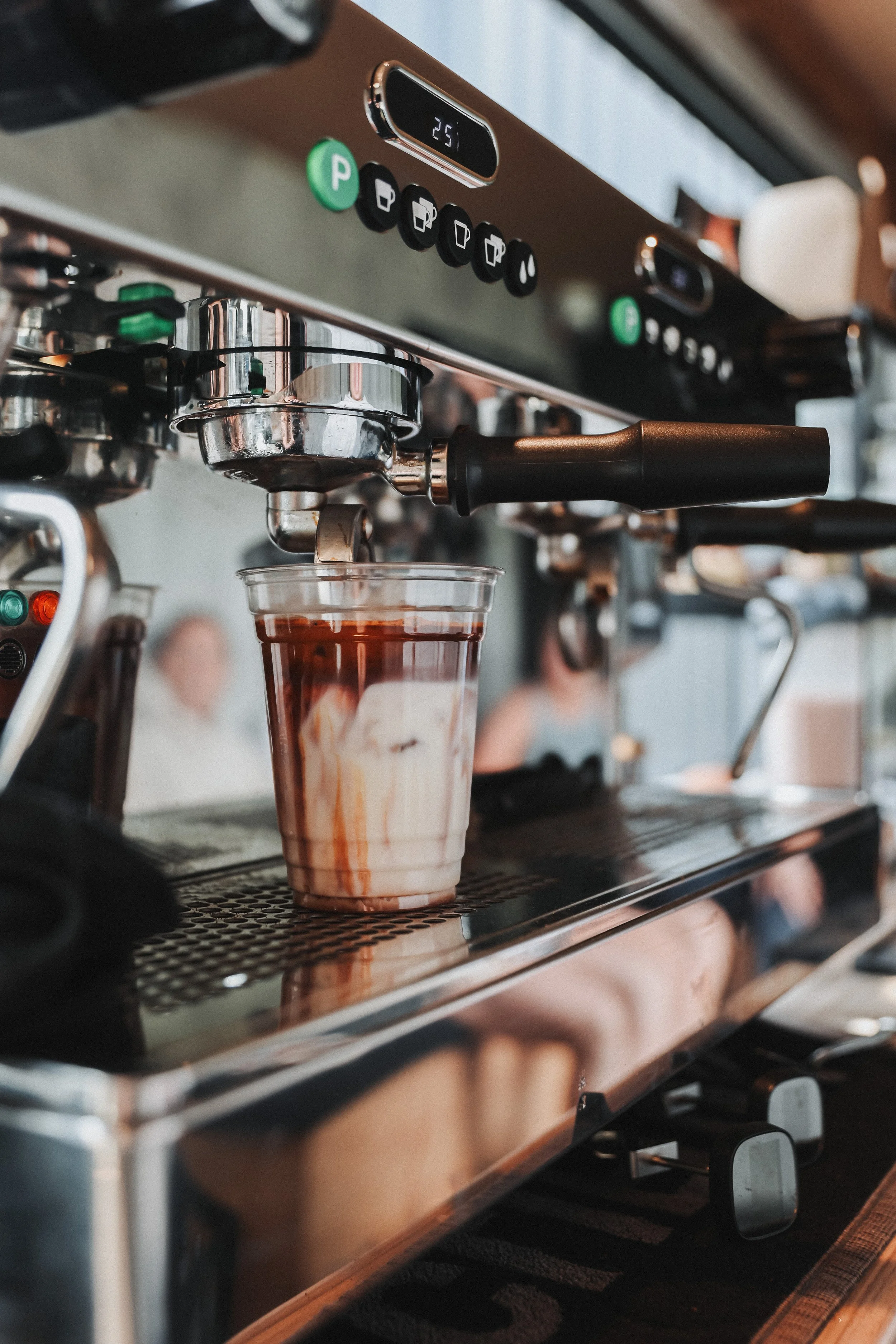 A close-up image of a professional espresso machine brewing a coffee drink, with a clear plastic cup underneath collecting the coffee, showing a layered cream and chocolate drink.