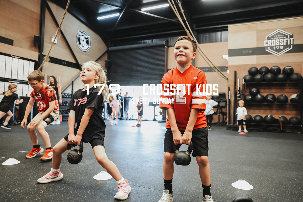 Children participating in a fitness class at CrossFit Kids gym, lifting kettlebells in a spacious, industrial-style gym with black rubber flooring and equipment along the walls.