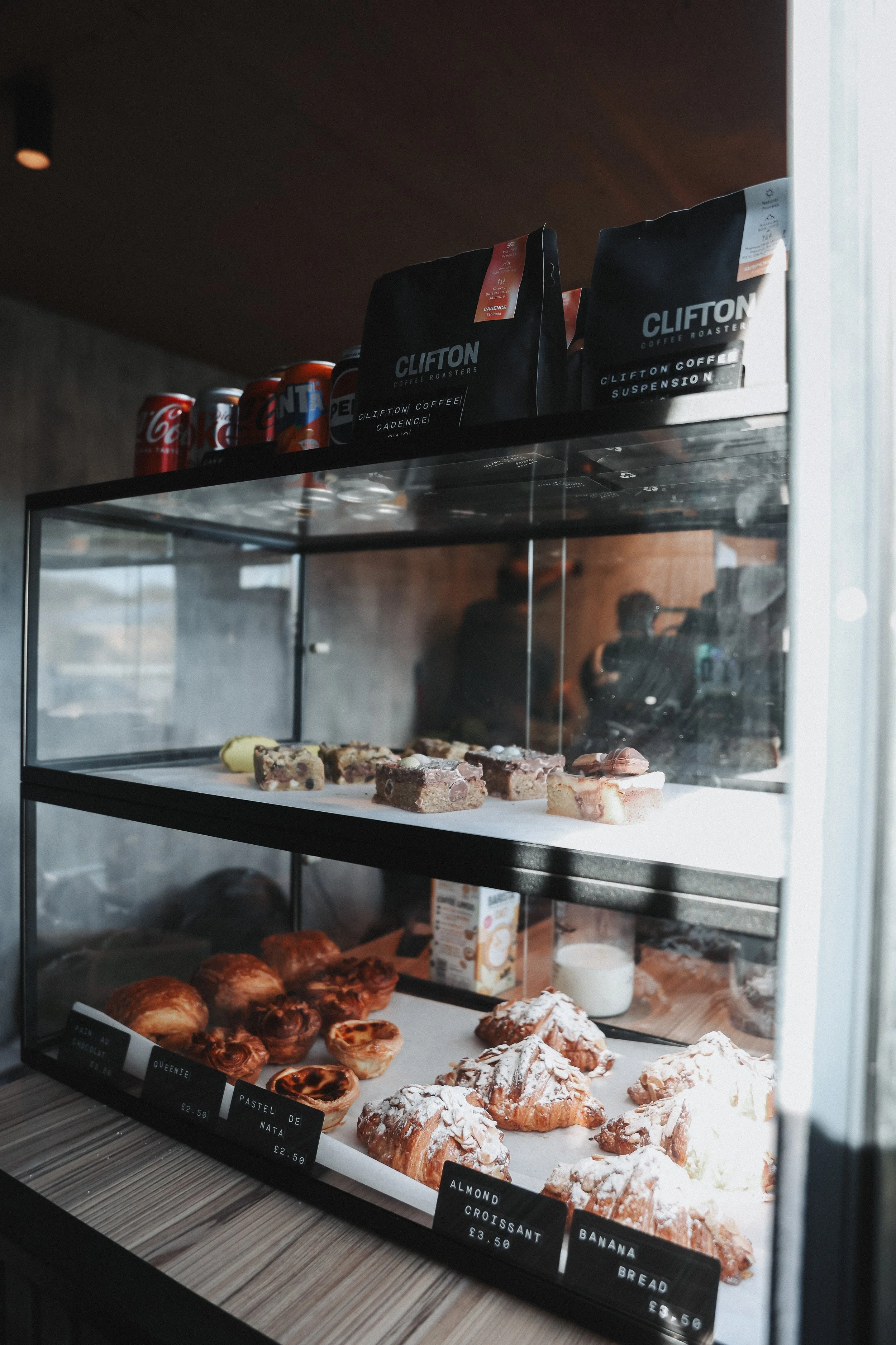Display case with assorted baked goods such as croissants and pastries, and packaged coffee and soda on the top shelf.