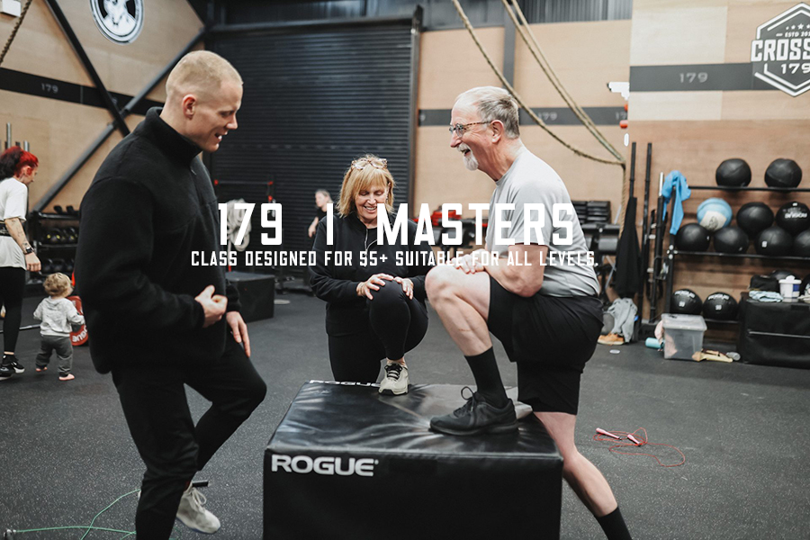 An older man doing a box jump exercise with a trainer and a woman observing in a gym with fitness equipment.