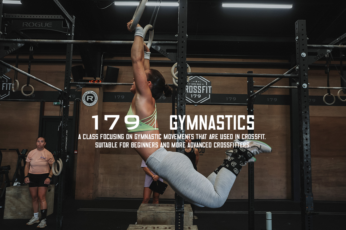 A woman doing gymnastic movements on gymnastic rings at CrossFit gym, with two women watching in the background.