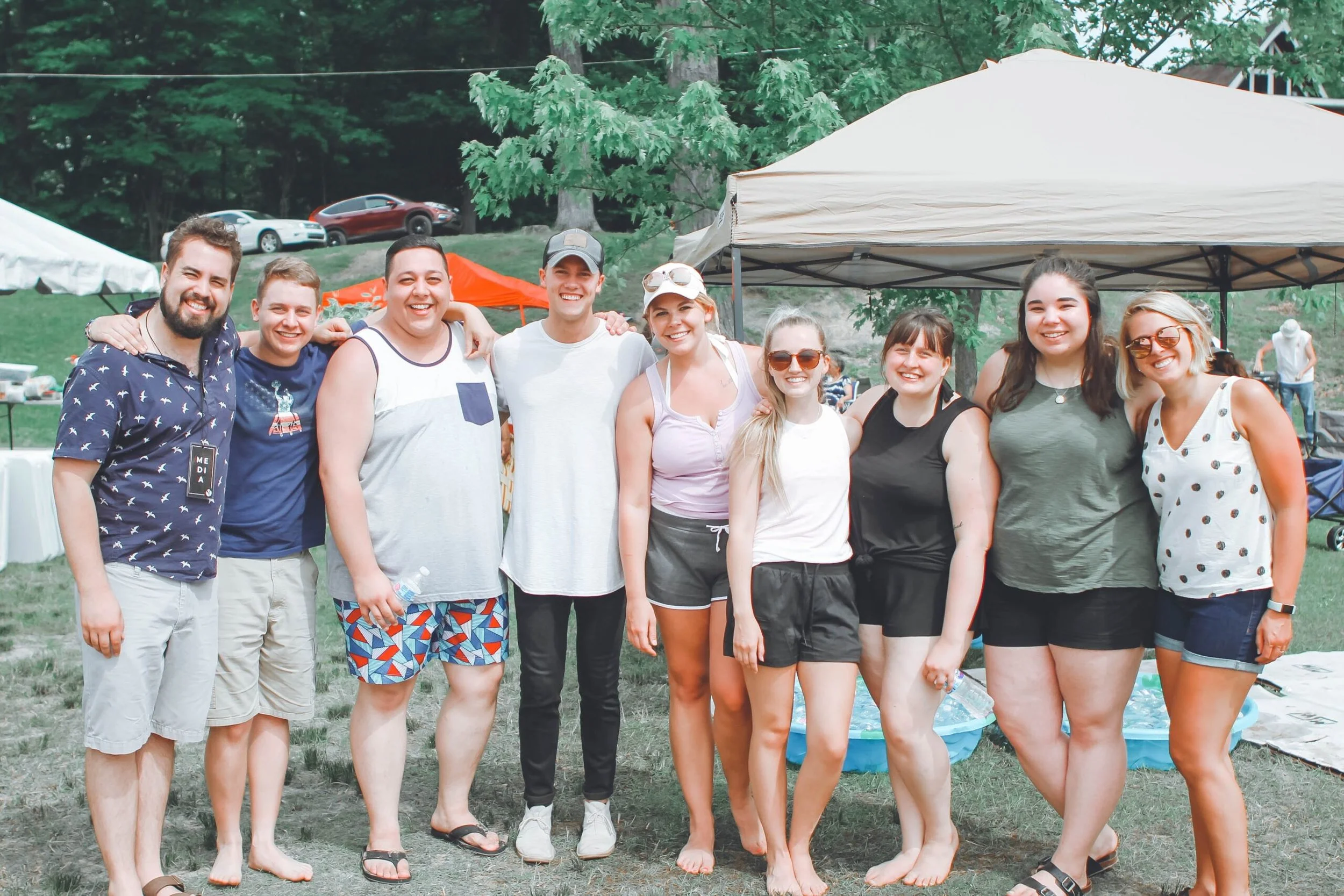 A group of ten friends smiling and standing closely together outdoors during daytime, with trees, cars, and tents in the background, on a grassy area.