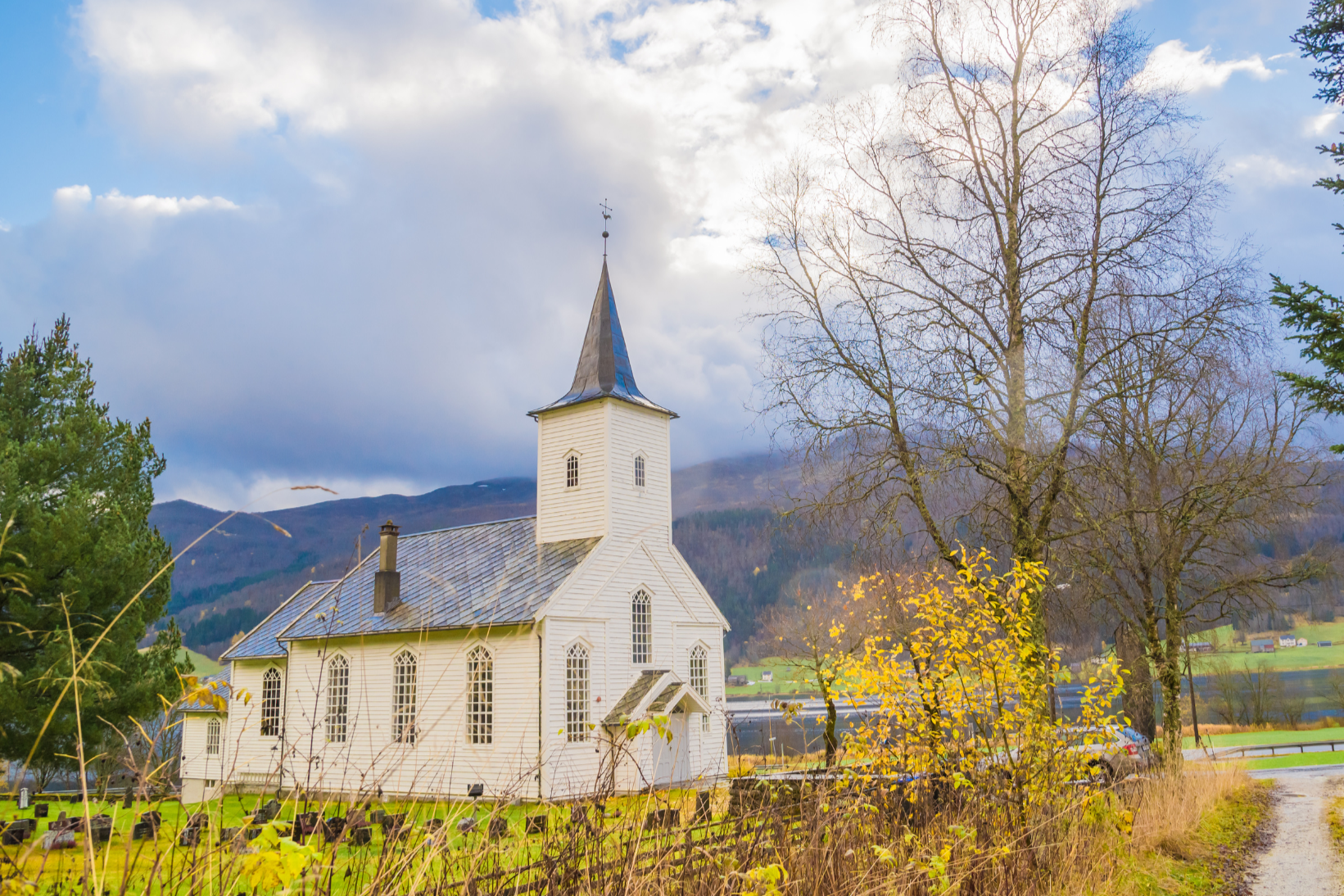 A white wooden church with a steeple, surrounded by trees with some yellow leaves, on a cloudy day with mountains in the background.