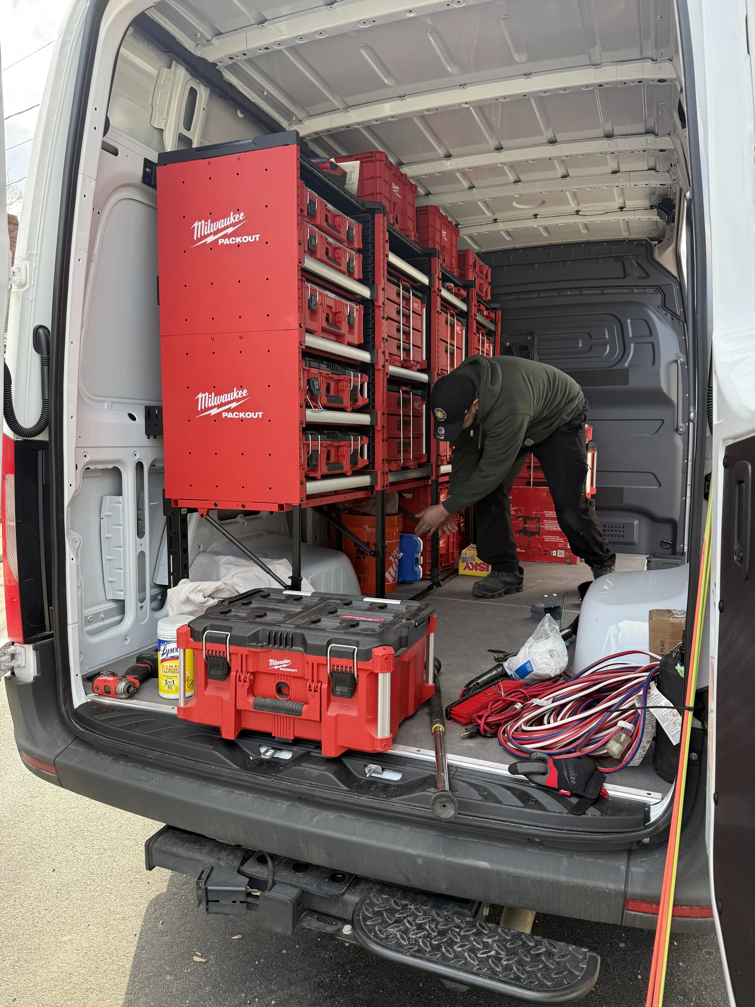 The back of a work van with its doors open, revealing organized tools, equipment, and supplies, including a large red Milwaukee tool storage unit, a trailer jack, and various cords and smaller tools on the floor. A person is bending over, working on something inside the van.