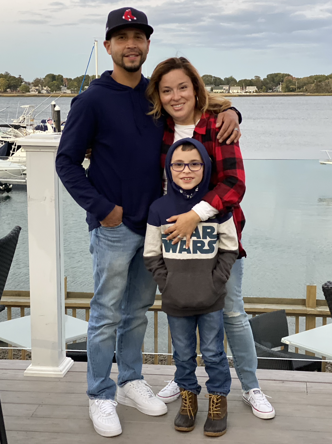 A family of three posing outdoors by a lake or river with boats in the background. The man is wearing a black baseball cap with a red logo, a navy hoodie, and light jeans. The woman is wearing a red and black plaid shirt over a white top and ripped jeans. The child, wearing glasses, a dark hoodie with a 'Star Wars' logo, and tan boots, is standing in front of them.