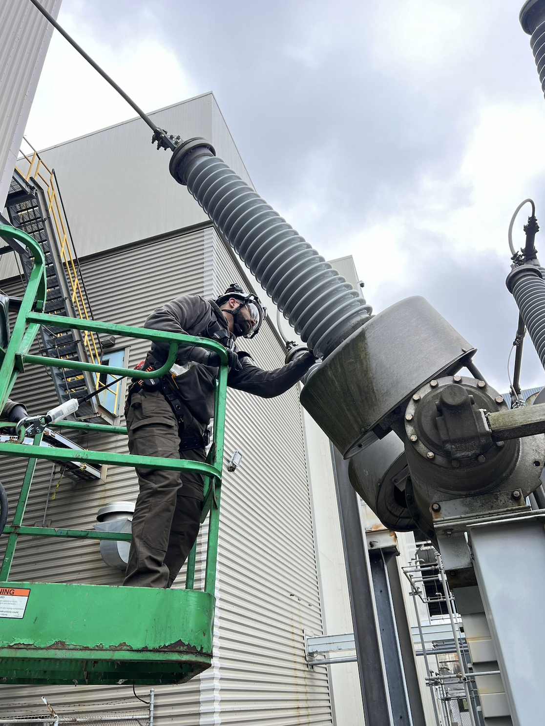 A technician working on electrical equipment on a green lift outside a large industrial building.
