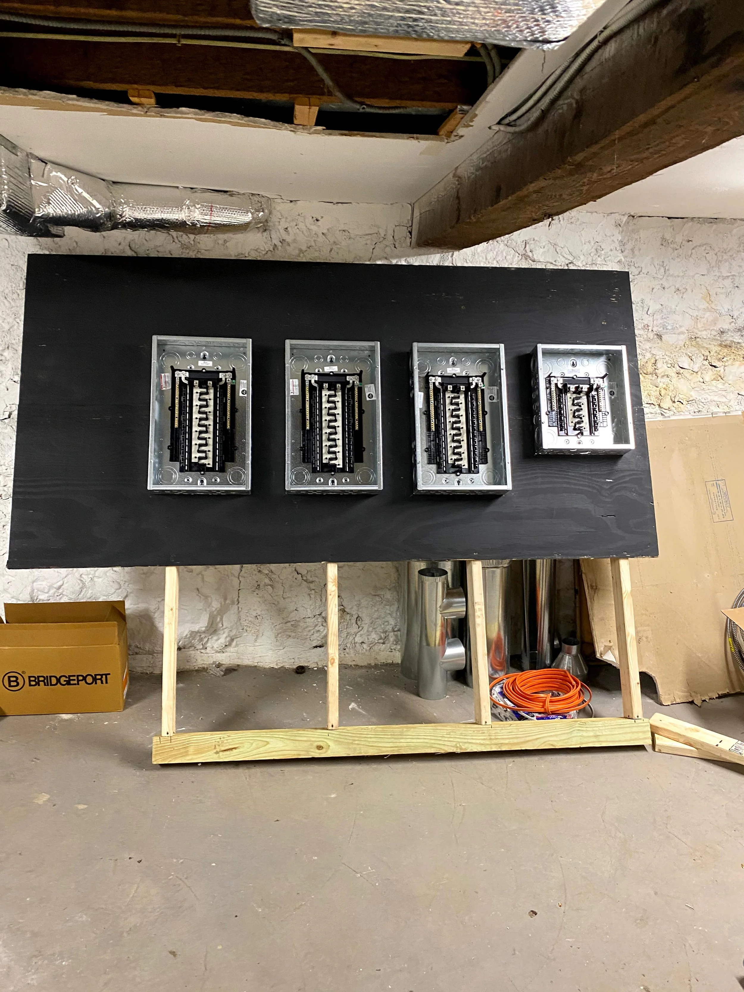 Construction area with electrical boxes mounted on a black board, wooden frame, toolbox, orange extension cord, and various construction materials on the floor.