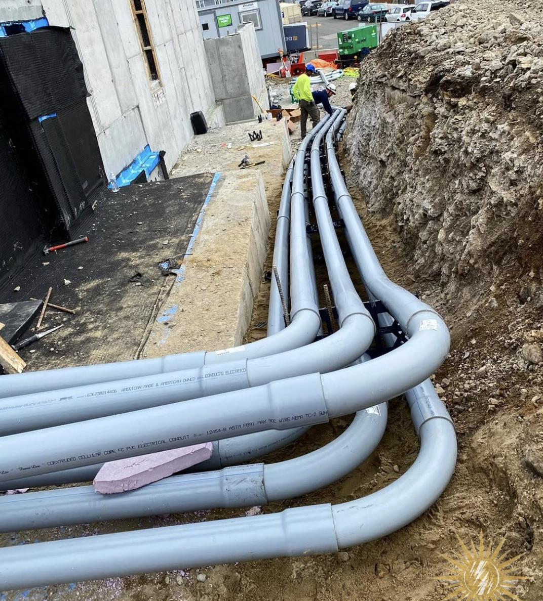 Construction workers install large gray electrical pipes underground next to a building foundation, with a rocky dirt trench on the right side and vehicles parked in the background.