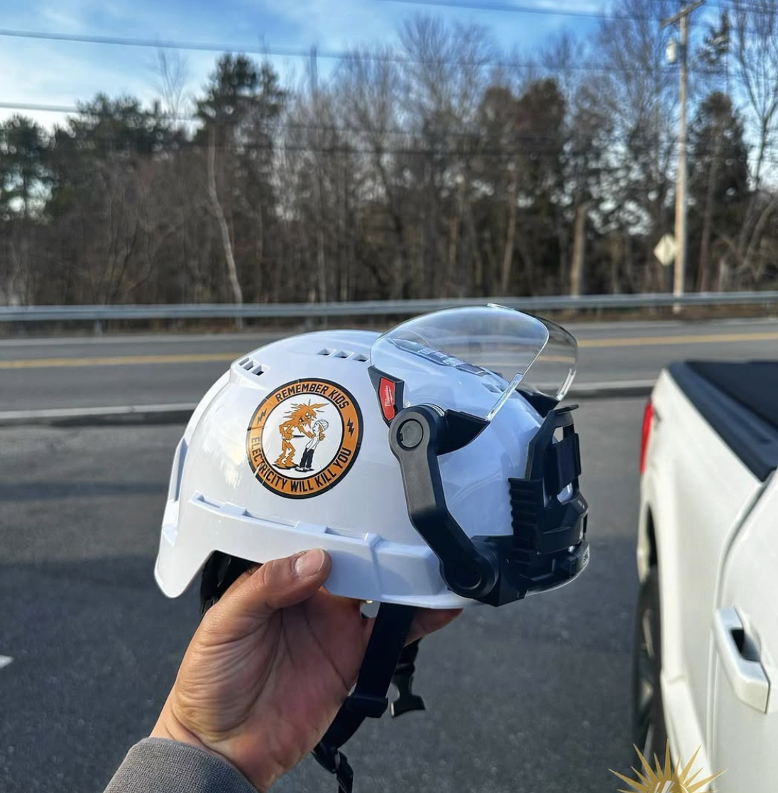 Person holding a white safety helmet with a sticker that reads 'Remember Kids Electricity Will Kill You' and has a cartoon image of a child and an electric current.