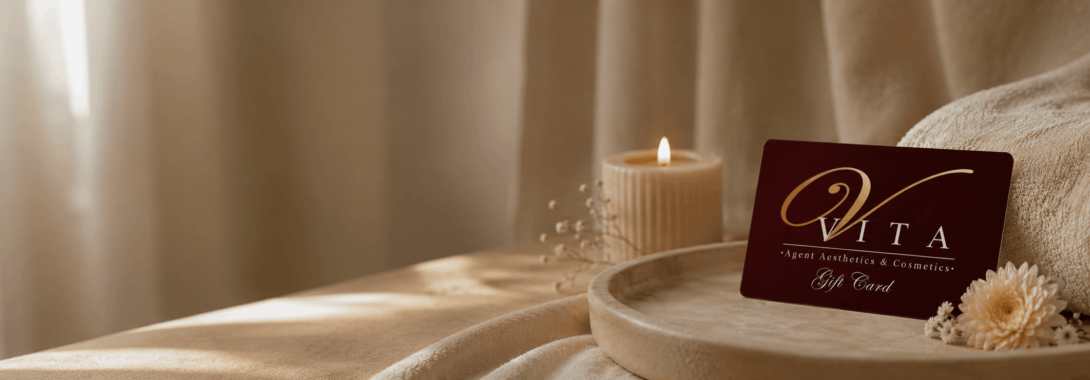 A gift card from VITA Agent Aesthetics & Cosmetics on a beige textured cloth with a lit candle, some dried flowers, and a beige towel, all arranged on a round beige tray for a spa or beauty salon setting.