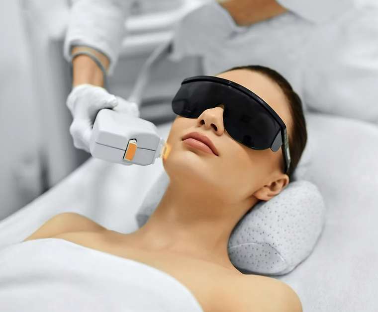 A woman lying down with protective eyewear while a technician performs a laser skin treatment on her face in a medical setting.