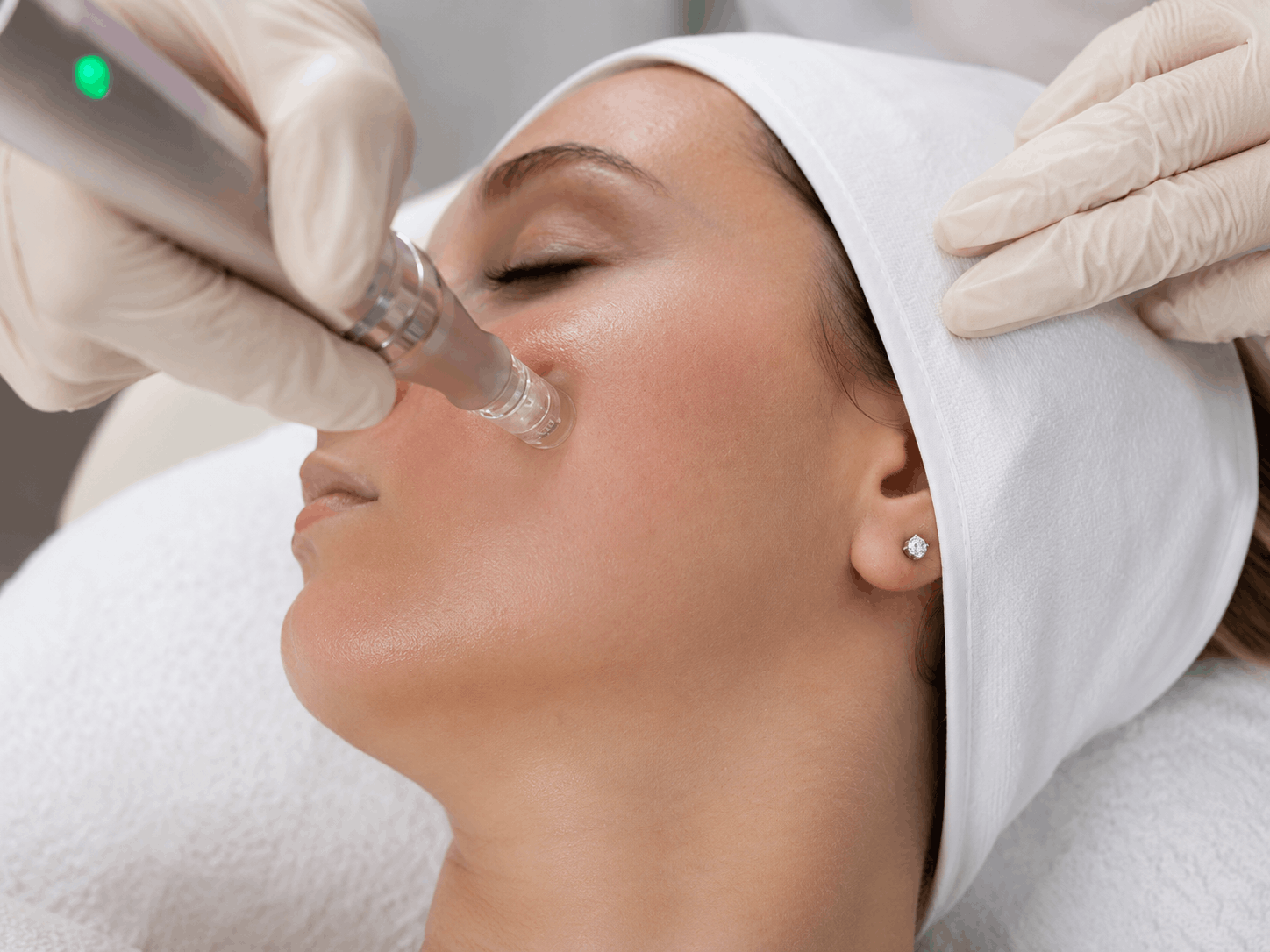 A woman receiving a cosmetic injection near her nose while lying down on a treatment bed, with a healthcare professional wearing pink gloves administering the procedure.