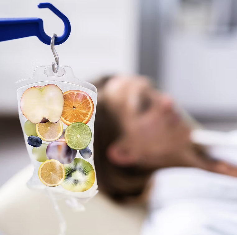 IV bag hanging in a hospital room filled with sliced fruit pieces including apple, orange, lemon, lime, blueberry, and kiwi, with a patient lying in the background.