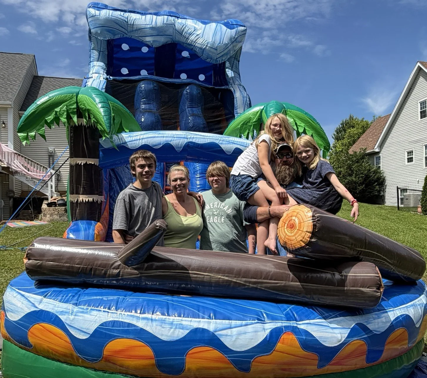 Family and friends enjoying a summer day on an inflatable water slide with a jungle and ocean theme in a backyard.