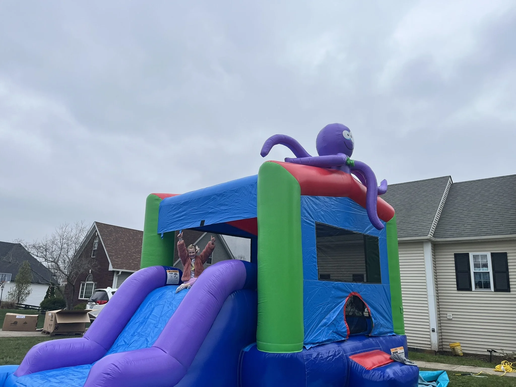 A colorful inflatable bounce house shaped like an octopus with a slide. A young girl is sitting at the top of the slide, smiling and waving. Houses and cars are visible in the background under a cloudy sky.